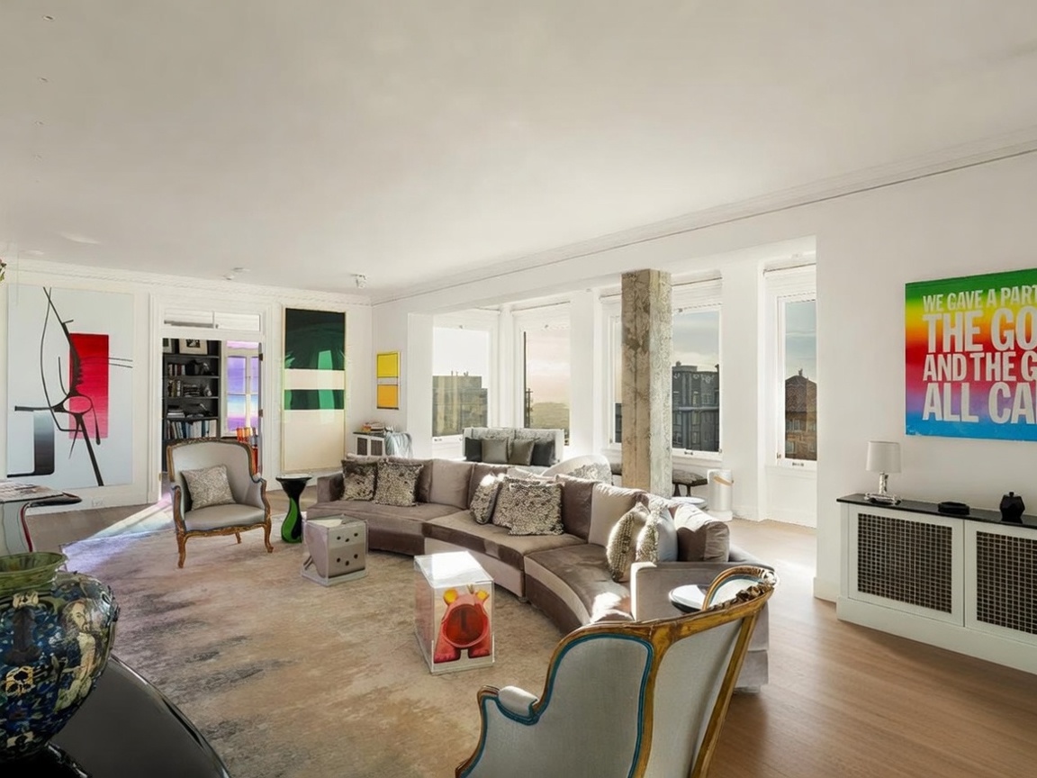 This expansive living room features a large, curved sectional sofa arranged on a textured area rug, creating a comfortable seating area. The space is decorated with eclectic art pieces, including a vibrant, text-based wall hanging and a tall abstract painting, while large windows provide ample natural light and city views. A central concrete pillar adds an industrial touch to the otherwise sophisticated and art-forward interior design.