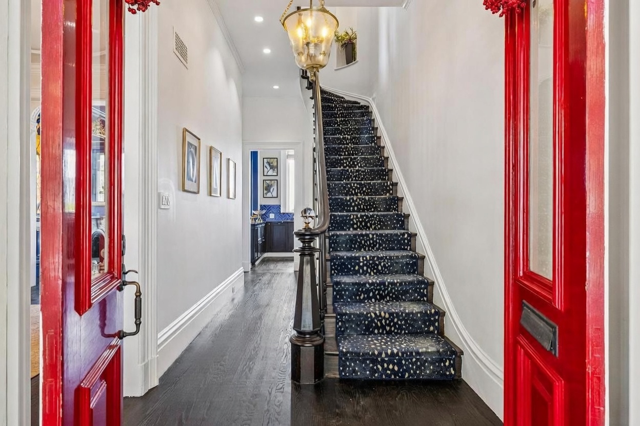This inviting entryway features a striking red door that opens into a bright hallway with dark hardwood floors. A staircase with a patterned runner leads to the upper level, complemented by a classic hanging lantern fixture. The perspective is from the doorway looking inward, showcasing a clean, elegant transition space that leads toward a glimpse of a blue-tiled room in the background.