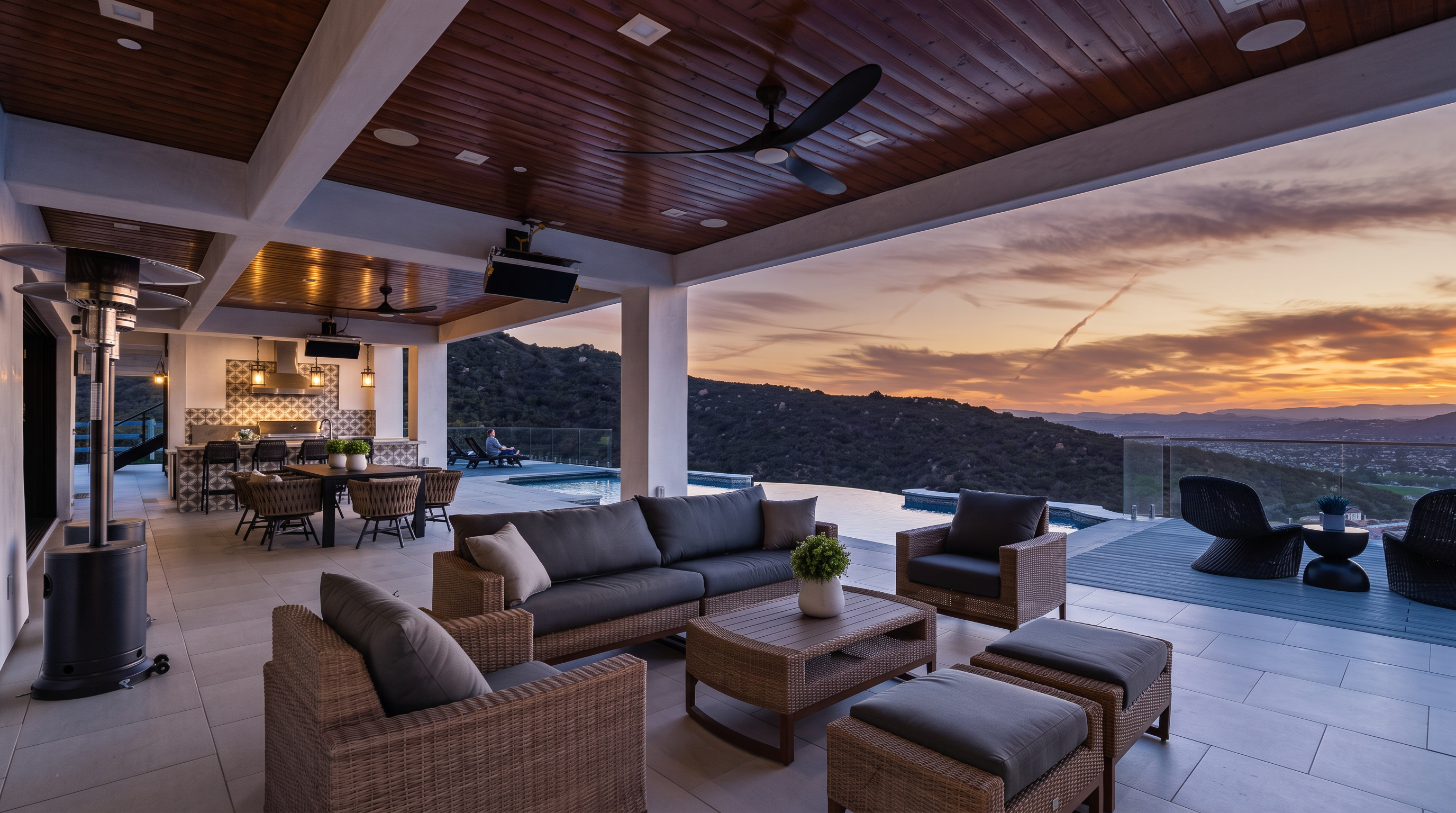 This luxurious covered outdoor living space features premium wicker furniture, a modern coffee table, and comfortable seating, all positioned to overlook an infinity-edge pool and sweeping mountain views. The area includes a well-equipped outdoor kitchen with high-end appliances, making it an ideal setting for al fresco dining and sophisticated entertaining. The combination of the warm wooden ceiling, clean tile flooring, and natural vistas creates an inviting and upscale atmosphere.