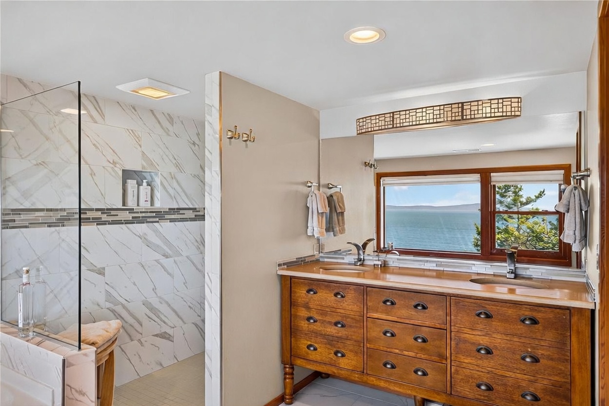 This primary bathroom features a dual-sink vanity with a warm-toned wood cabinet and a large mirror reflecting a scenic water view through a wide window. To the left, a walk-in shower is enclosed by glass and finished with light-colored patterned tile and a decorative mosaic accent strip. The space is well-lit with recessed lighting and offers a serene, spa-like atmosphere with a direct connection to the outdoors.