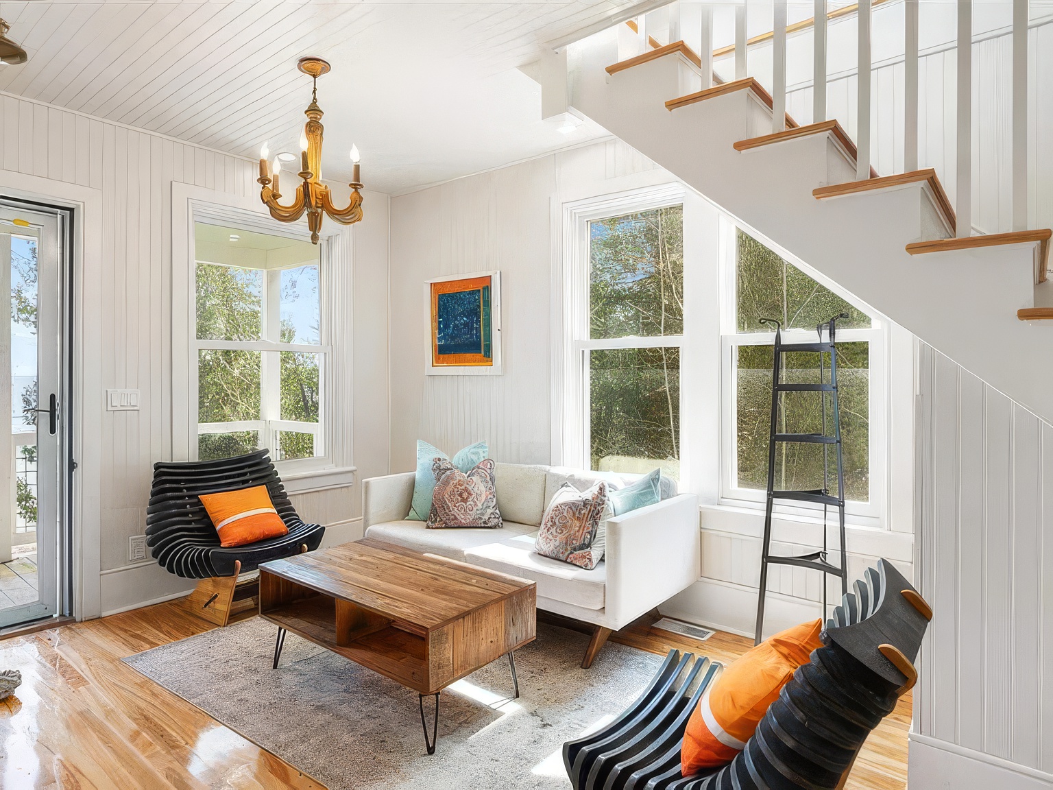 This bright living room features white vertical wood paneling and light-toned hardwood flooring, creating a clean, coastal-inspired aesthetic. The space is furnished with a white sofa, two unique slatted black chairs with orange accent pillows, and a rustic wood coffee table centered on a textured area rug. A staircase with white railings ascends along the right side, while large windows provide ample natural light and views of the surrounding greenery.