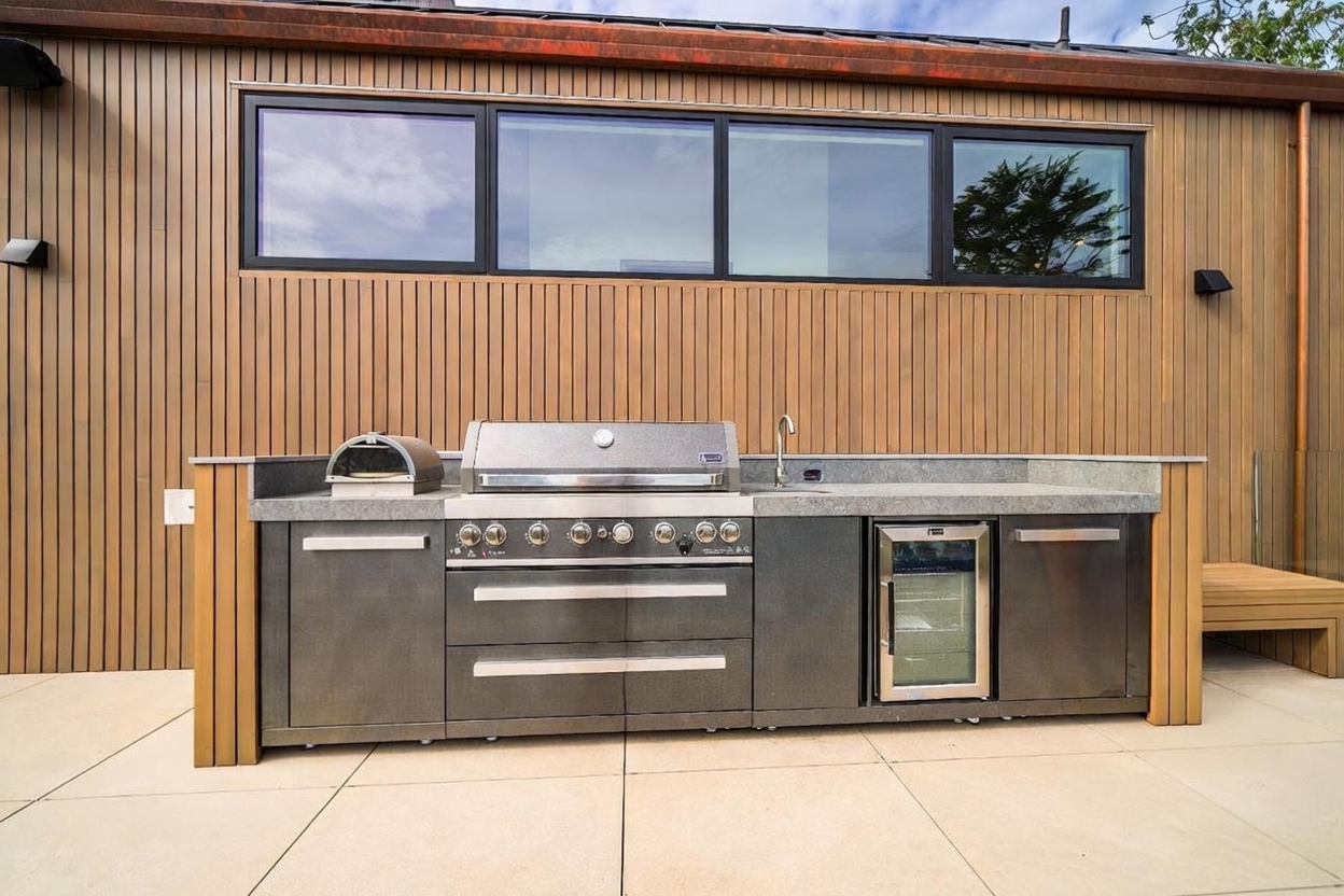 This outdoor kitchen area features a sleek, modern design with vertical wood-slat siding and a stone countertop. The setup includes a large stainless steel grill, a built-in pizza oven, a sink, and an under-counter beverage refrigerator. Large windows above the cooking station provide a view into the home, creating a seamless transition between indoor and outdoor living spaces.