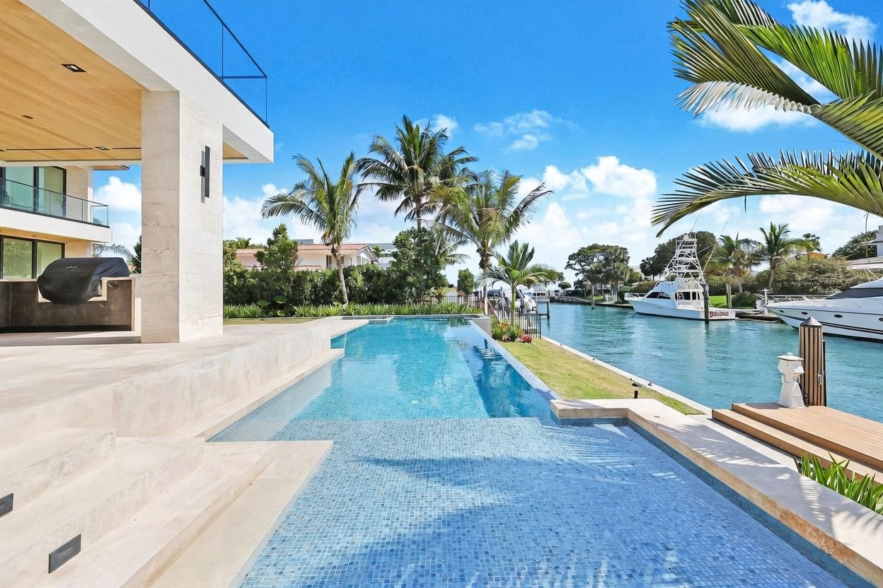 This luxurious waterfront property features a sleek, rectangular infinity-edge pool with blue mosaic tiling that seamlessly blends into the canal view. The expansive patio area is finished in light-toned stone, providing a clean, modern aesthetic that complements the contemporary architecture of the home. In the background, palm trees and docked boats enhance the serene, tropical atmosphere of this high-end residential setting.