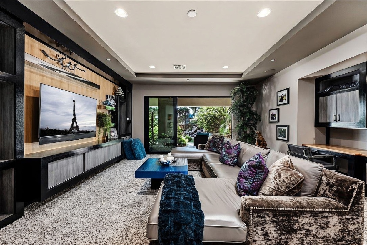 This spacious living room features a large, plush sectional sofa adorned with patterned throw pillows and a deep blue knit throw. A sleek, dark-toned media console with light wood paneling serves as the focal point, housing a large television displaying an image of the Eiffel Tower. Large sliding glass doors provide a seamless transition to the outdoor patio, while recessed lighting and a neutral color palette create a sophisticated and inviting atmosphere.