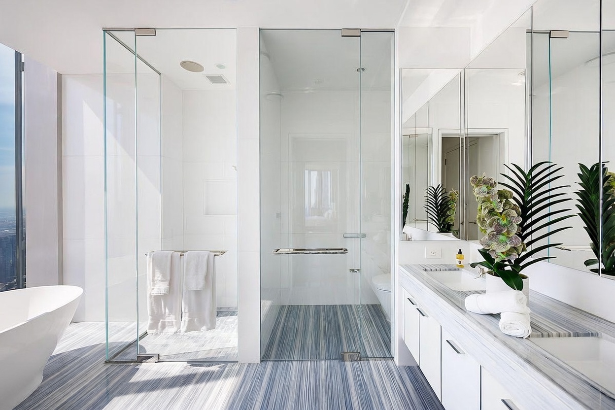 This modern primary bathroom features a sleek, minimalist aesthetic with a striking blue and white linear-patterned stone floor. The space includes a glass-enclosed walk-in shower, a freestanding soaking tub, and a long vanity with dual sinks and large mirrors. The bright, clean design is accented by a decorative plant on the vanity, creating a serene and luxurious atmosphere.