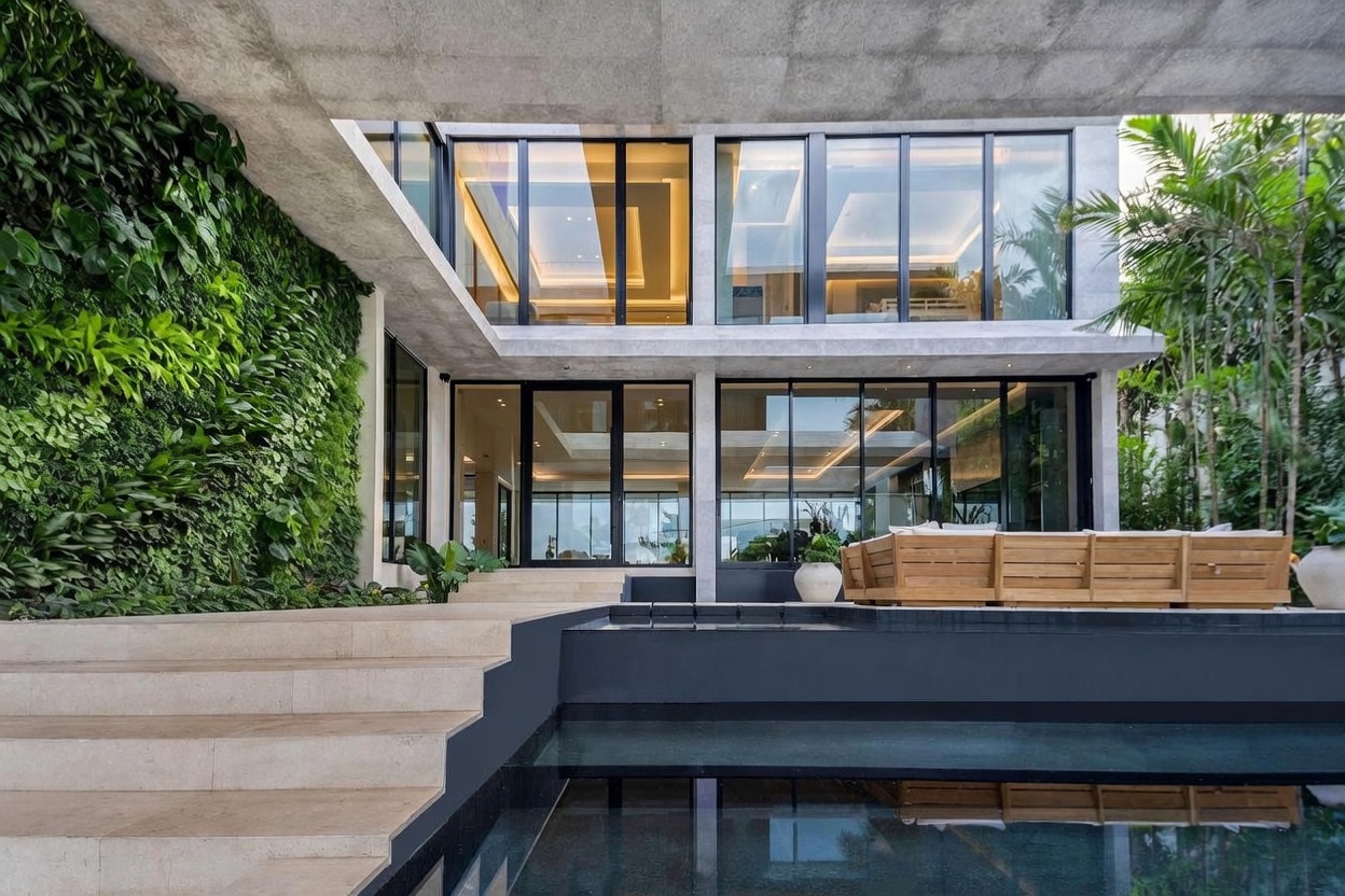 This contemporary residence features a striking architectural design with a prominent living green wall and a sleek, dark-tiled pool in the foreground. The structure utilizes extensive floor-to-ceiling glass panels that blur the line between indoor and outdoor living spaces, complemented by a concrete overhang and light-toned stone steps. A wooden outdoor sofa sits on the patio, creating a serene and luxurious atmosphere perfect for relaxation.