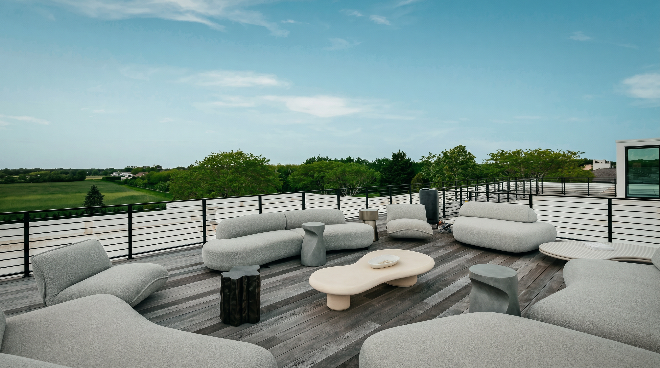 This expansive rooftop deck features a modern, minimalist aesthetic with curved, light-toned outdoor seating arranged around organic-shaped coffee tables. The space is framed by a sleek black metal railing that overlooks a lush, green landscape under a clear blue sky. The weathered wood decking provides a natural texture that complements the contemporary furniture, creating an inviting atmosphere for outdoor entertaining.