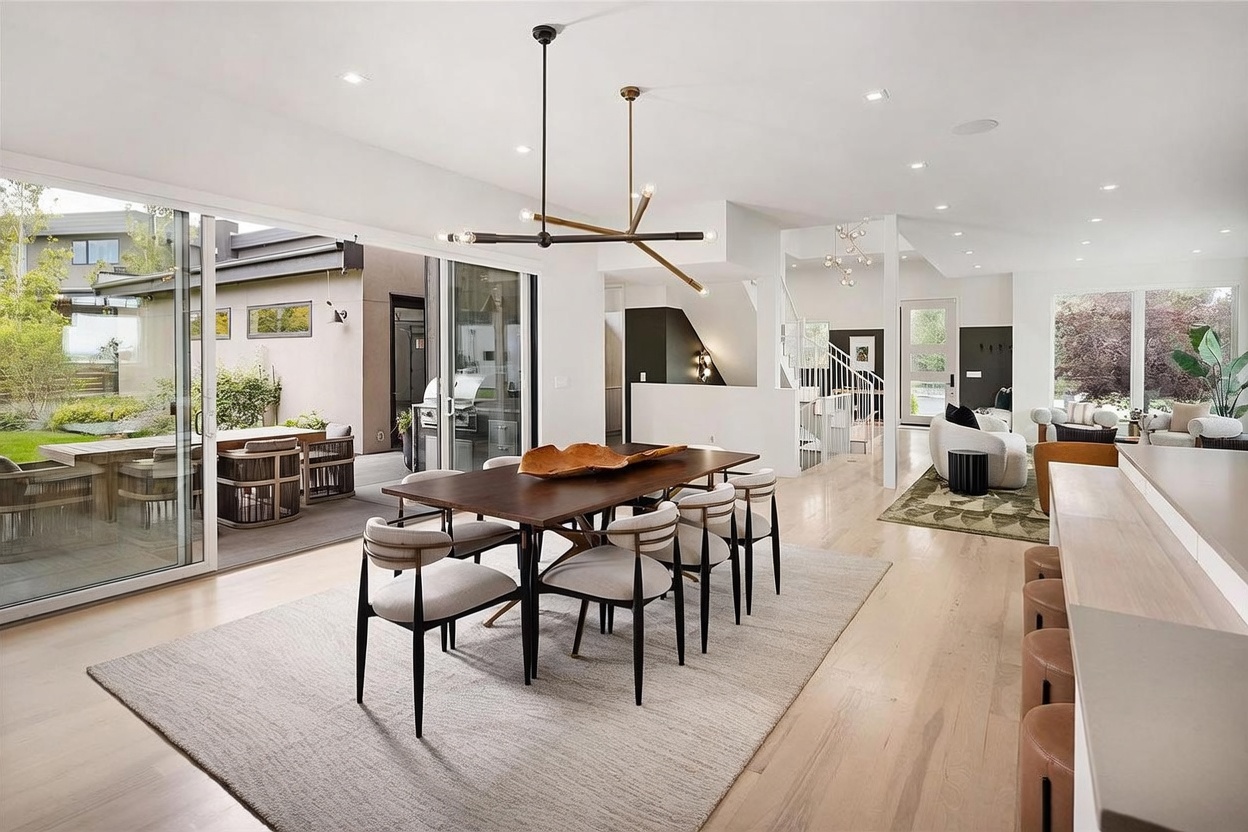 This open-concept dining area features a dark wood table surrounded by six upholstered chairs, set atop a textured area rug. A modern, multi-armed light fixture hangs above the table, while large sliding glass doors provide a seamless transition to the outdoor patio. The space flows into a bright living area and kitchen, showcasing light wood flooring and a clean, contemporary aesthetic.