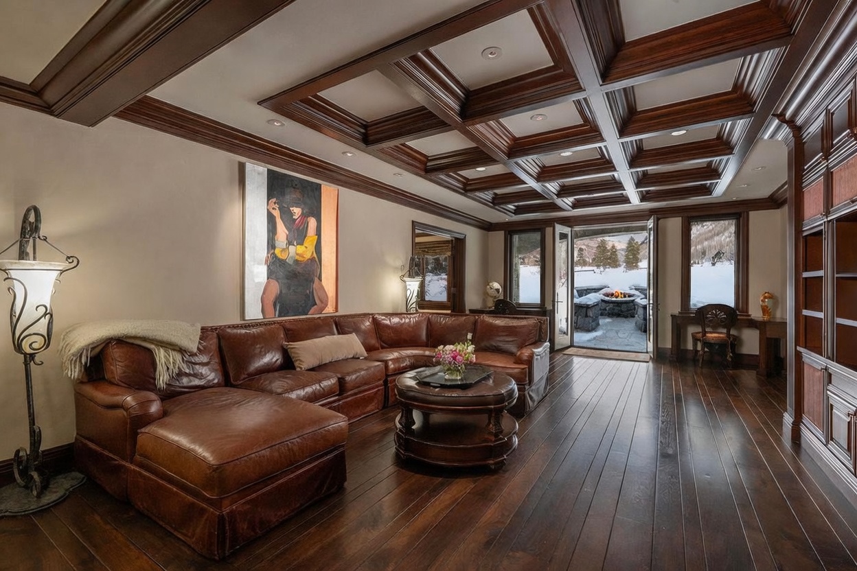This sophisticated living room features a large, L-shaped leather sectional sofa arranged around a circular leather ottoman, set against a backdrop of dark wood flooring and a coffered ceiling. A striking piece of modern art hangs on the wall, while double doors open to a snowy outdoor patio area with a stone fire pit. The room exudes a warm, luxurious mountain aesthetic with rich wood tones and elegant lighting.