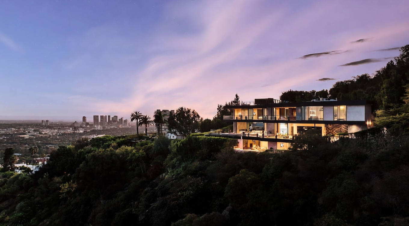 This stunning aerial shot captures a modern, multi-level luxury residence perched on a lush hillside overlooking a sprawling city skyline at dusk. The home features expansive glass walls, sleek architectural lines, and illuminated terraces that create a sophisticated, high-end aesthetic. The dramatic purple and orange sunset sky provides a cinematic backdrop, emphasizing the property's exclusive and elevated position.