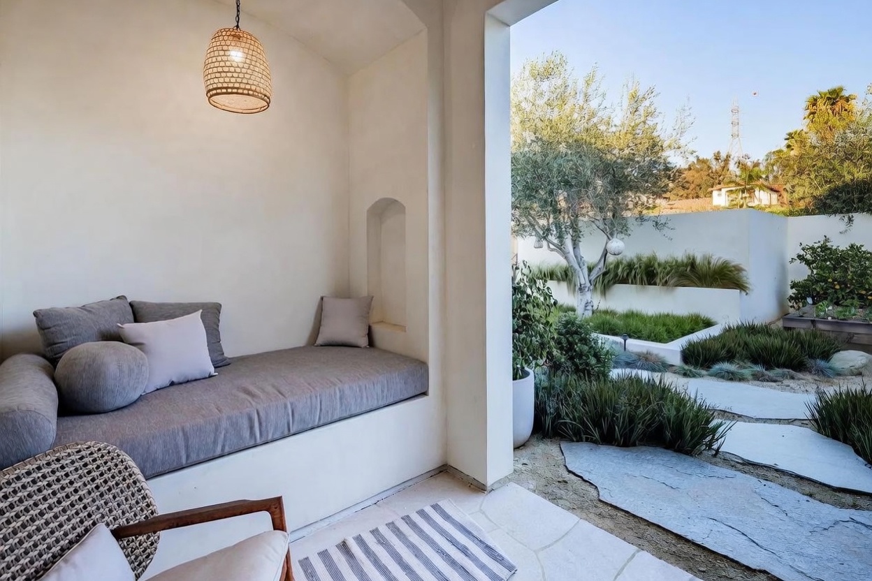 This serene outdoor living space features a built-in bench with plush gray cushions, nestled within a recessed alcove under a covered patio. A woven pendant light hangs above the seating area, which transitions seamlessly to a landscaped garden featuring stone pavers, ornamental grasses, and a mature olive tree. The overall aesthetic is minimalist and Mediterranean-inspired, offering a tranquil retreat with a clear view of the surrounding yard.