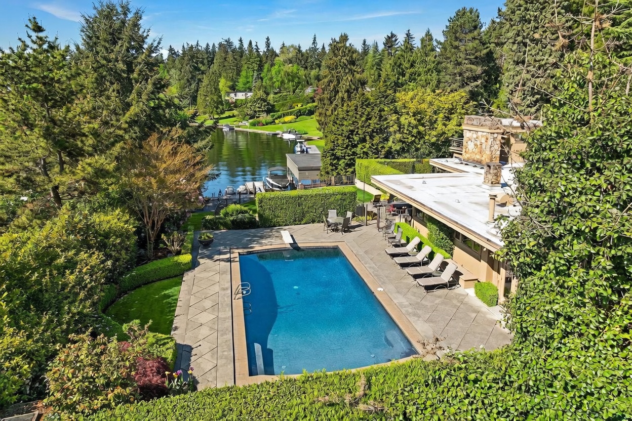 This high-angle aerial view captures a luxurious waterfront property featuring a rectangular swimming pool with a diving board set into a spacious stone patio. The home includes a flat roof and a prominent stone-clad chimney breast, surrounded by lush, mature landscaping and tall trees. Beyond the manicured hedges, a private dock extends into the calm lake, creating a serene and private outdoor living environment.