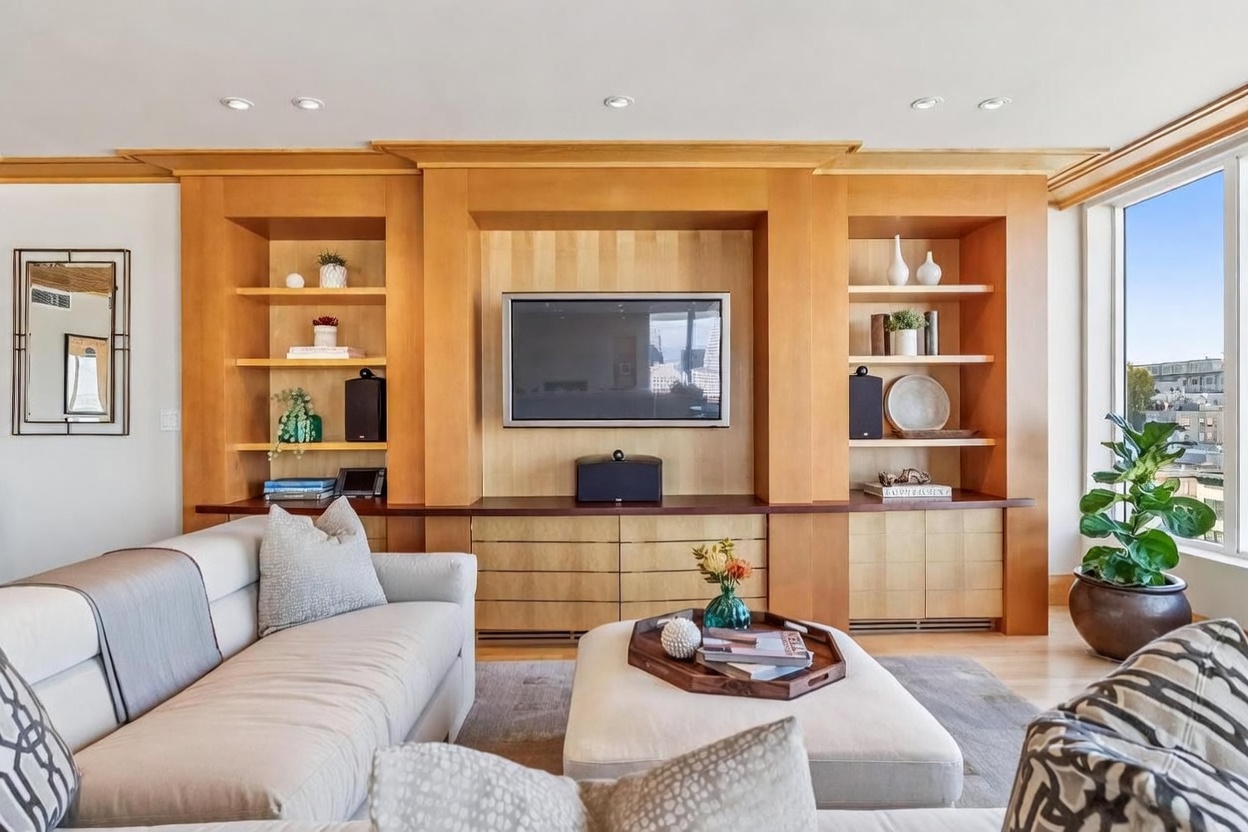 This bright living room features a large, custom light wood media wall with integrated shelving and cabinetry surrounding a central television. A plush, light-colored sectional sofa and a matching ottoman with a wooden tray create a comfortable seating area, while a large window to the right provides natural light and a view of the cityscape. The space is decorated with tasteful accents, including a potted plant and a framed mirror, offering a clean and sophisticated aesthetic.