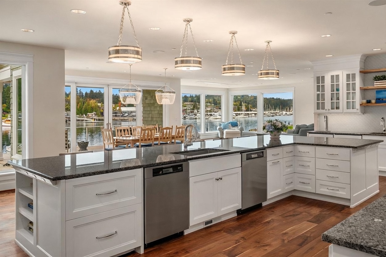 This bright, open-concept kitchen features a large L-shaped island with a dark stone countertop and white cabinetry, complemented by warm wood flooring. The space is illuminated by multiple pendant lights and offers expansive views of the water and marina through large glass doors and windows. The design creates a seamless transition between the functional kitchen area and the adjacent dining space, emphasizing a light and airy atmosphere.