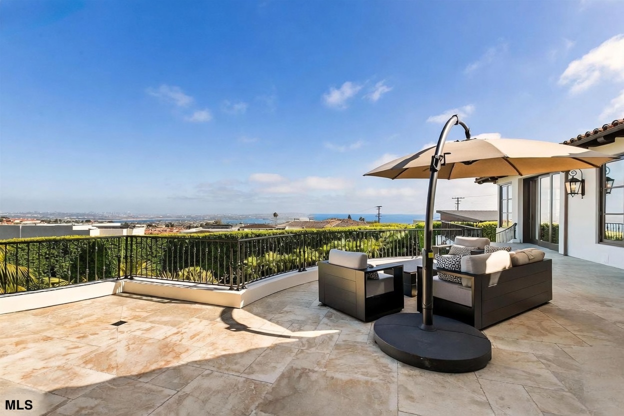 This expansive stone-paved terrace offers a luxurious outdoor living space with a panoramic view of the coastline and city below. The area is furnished with comfortable lounge seating shaded by a large cantilevered umbrella, creating an ideal spot for relaxation. A decorative black metal railing borders the patio, which is framed by manicured hedges and overlooks a scenic landscape.