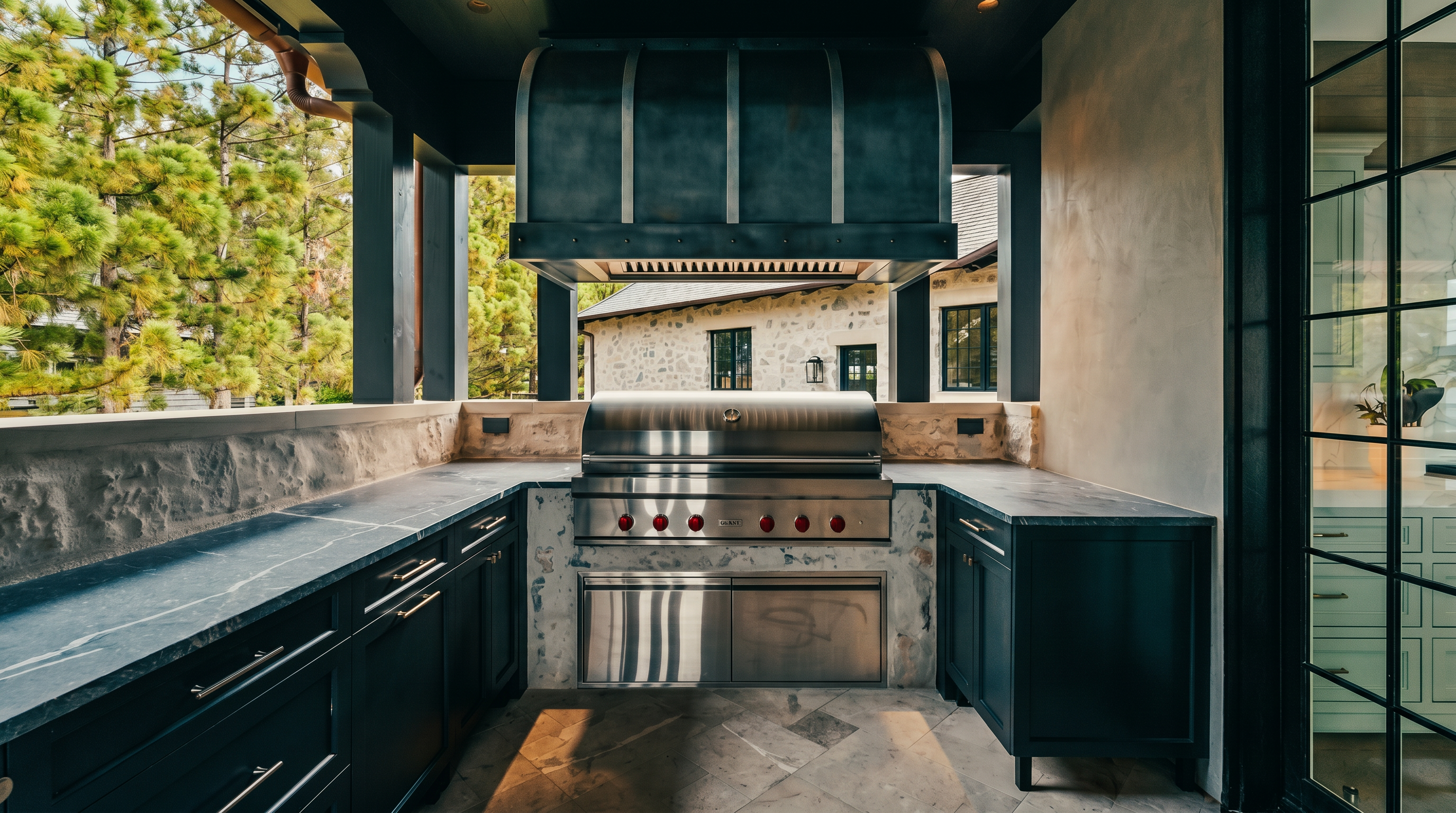 This sophisticated outdoor kitchen area features a professional-grade stainless steel grill set into a stone-clad base with dark cabinetry and stone countertops. The space is covered by a dark-painted ceiling with a mounted fan, creating a sheltered and elegant cooking environment. Large windows to the right provide a view into the home, while the open design overlooks a stone-walled exterior area.