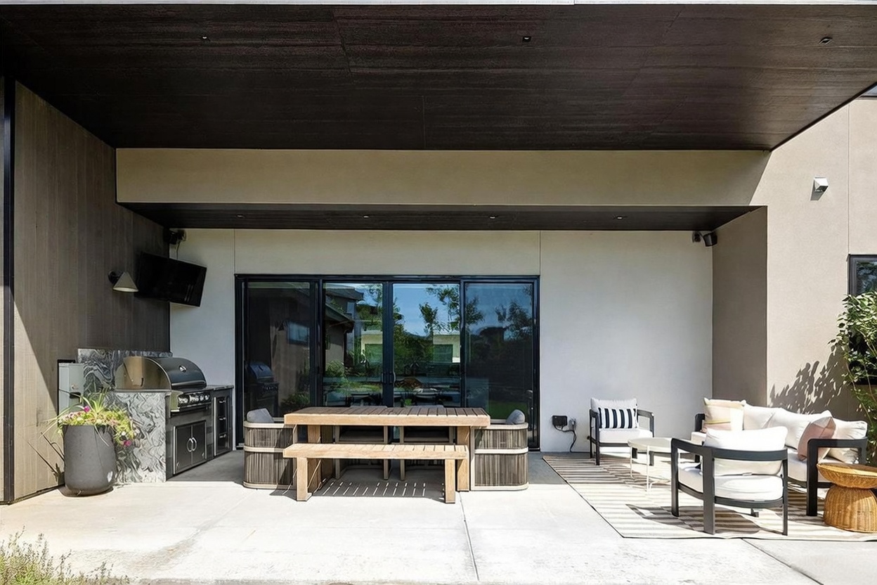 This modern covered patio features a built-in outdoor kitchen with a stainless steel grill and stone-clad counter, adjacent to a dining area with a wooden table and bench seating. A separate lounge area sits on a striped rug, complete with contemporary outdoor chairs and a sofa. The space is defined by a dark, flat ceiling and large glass sliding doors that connect the indoor and outdoor living areas.