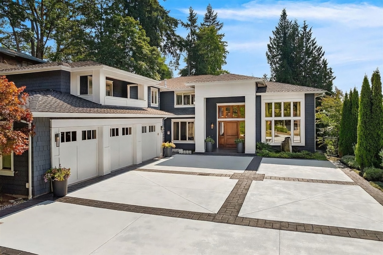 This striking two-story residence features a modern aesthetic with dark gray shingle siding contrasted by crisp white trim and a prominent, light-colored entryway structure. The expansive concrete driveway is segmented by decorative brick-style pavers, leading to a three-car garage on the left and a grand front entrance. Large, multi-pane windows allow for natural light, while the surrounding mature trees and manicured landscaping enhance the home's curb appeal.