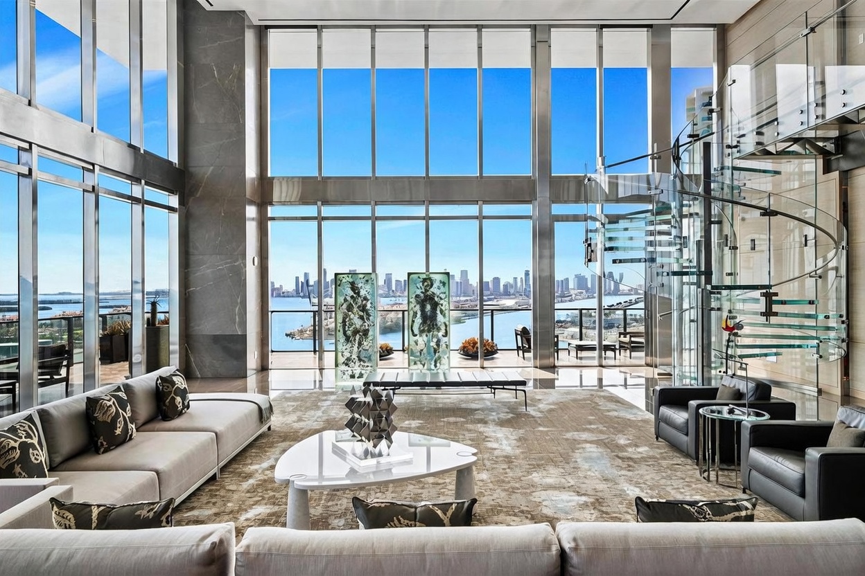 This expansive, double-height living room features dramatic floor-to-ceiling windows that frame a panoramic view of the city skyline and waterfront. The space is anchored by a large, neutral-toned area rug, a contemporary white triangular coffee table, and a sleek glass spiral staircase that adds a sculptural element to the room. Two striking, translucent art installations stand prominently before the glass, enhancing the sophisticated and airy atmosphere of this luxury residence.
