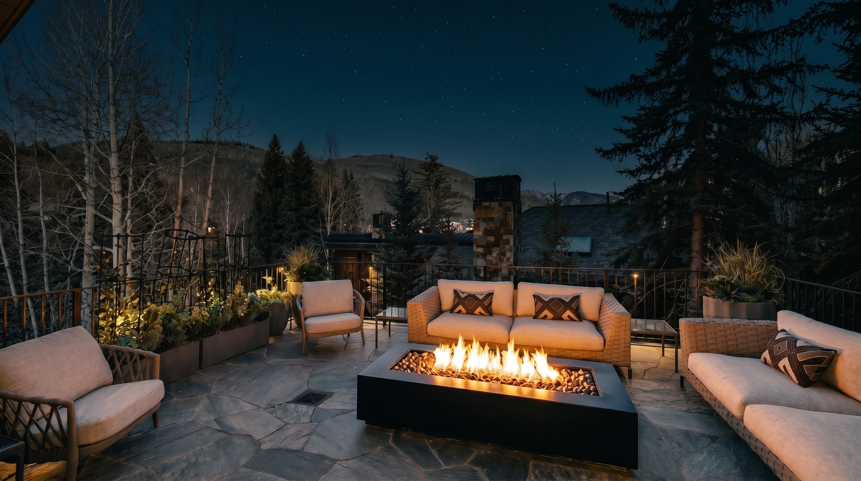 This expansive stone-paved terrace offers a sophisticated outdoor living space, featuring a modern rectangular fire pit as its centerpiece. The area is furnished with comfortable, neutral-toned seating, including a sofa and armchairs, and is bordered by metal railings and elevated planters with trellises. The setting provides a scenic, elevated view of the surrounding mountain landscape and evergreen trees, creating an inviting atmosphere for relaxation.