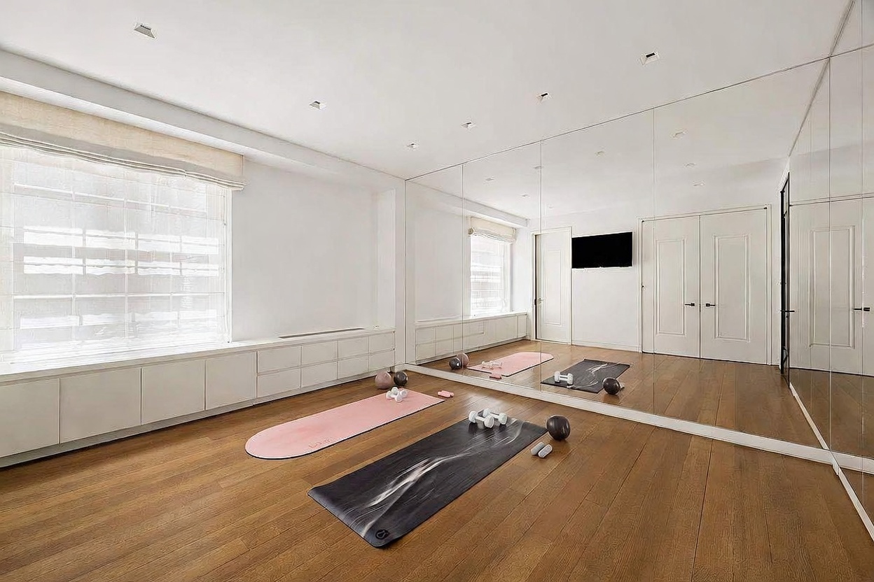 This bright and minimalist home gym features light wood flooring and a wall of floor-to-ceiling mirrors that enhance the sense of space. The room is equipped with yoga mats, small hand weights, and a wall-mounted television, while built-in cabinetry along the window wall provides convenient storage. Large windows with neutral shades allow for ample natural light, creating an airy and inviting atmosphere for exercise.