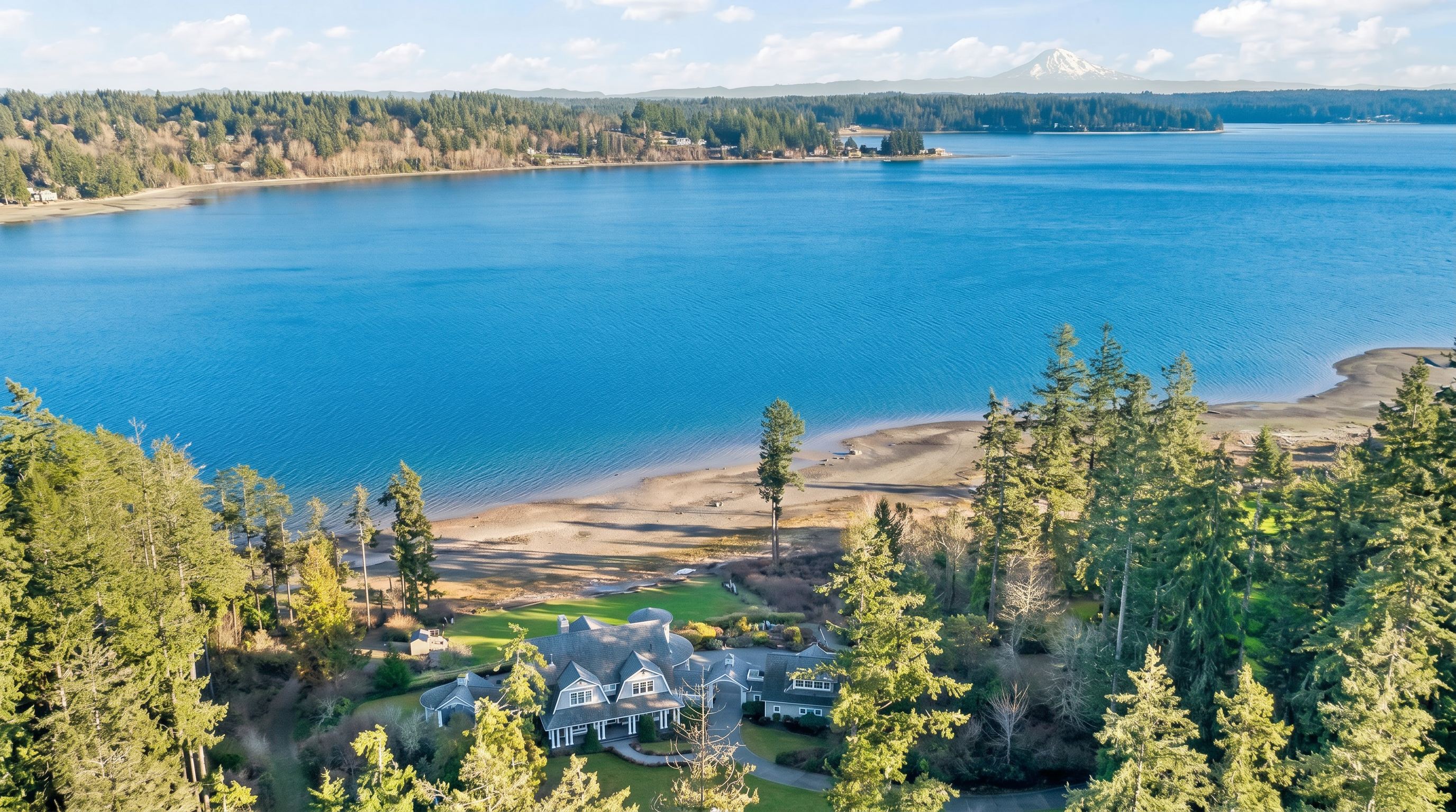 This high-angle aerial view captures a sprawling waterfront estate nestled within a dense forest, overlooking a vast blue bay with a prominent snow-capped mountain in the distance. The home features a complex, multi-gabled roofline and a circular driveway, surrounded by lush green lawns that transition into a natural sandy shoreline. The perspective emphasizes the property's secluded, serene location and its expansive connection to the surrounding natural landscape.