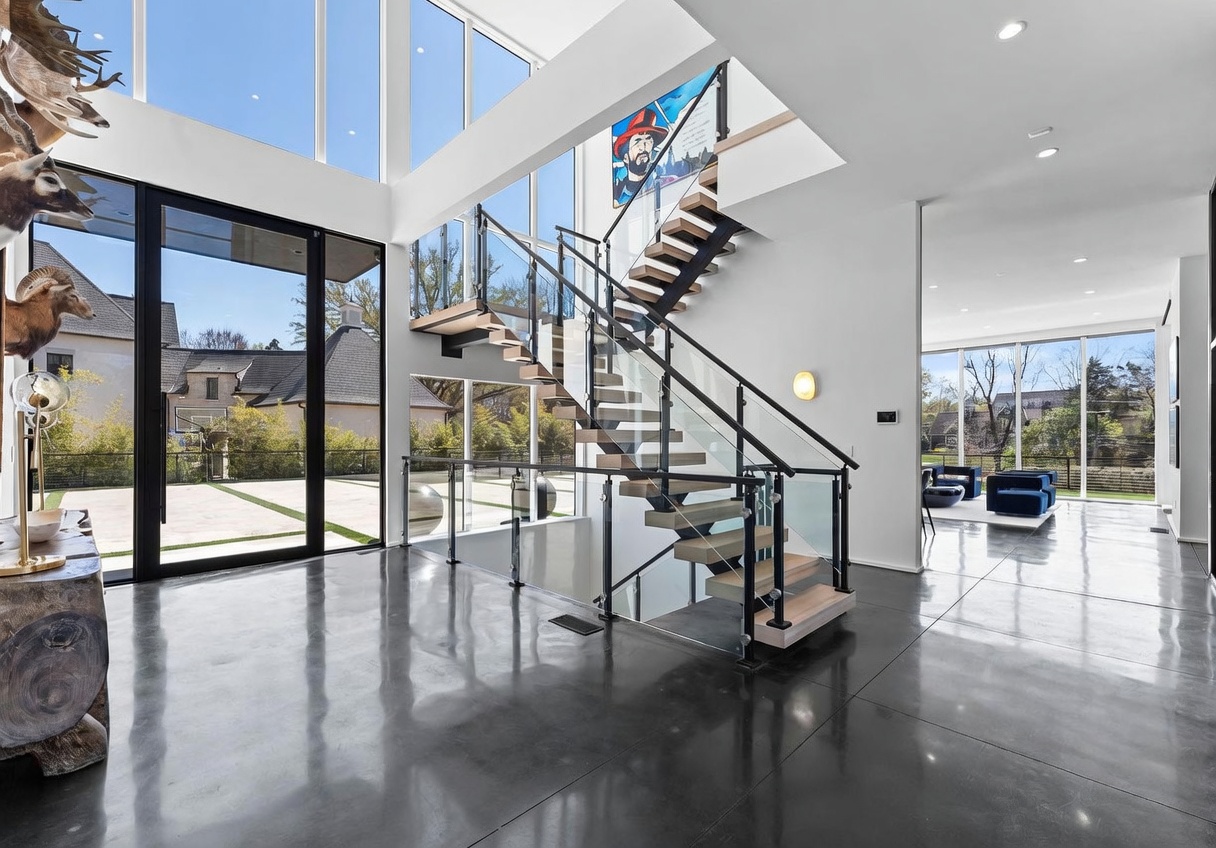 This dramatic entryway features a striking open-riser staircase with glass railings that serves as the focal point of the double-height space. The room is characterized by polished concrete floors and expansive glass walls that provide abundant natural light and views of the surrounding landscape. A large piece of wall art adds a vibrant pop of color to the minimalist, modern aesthetic.