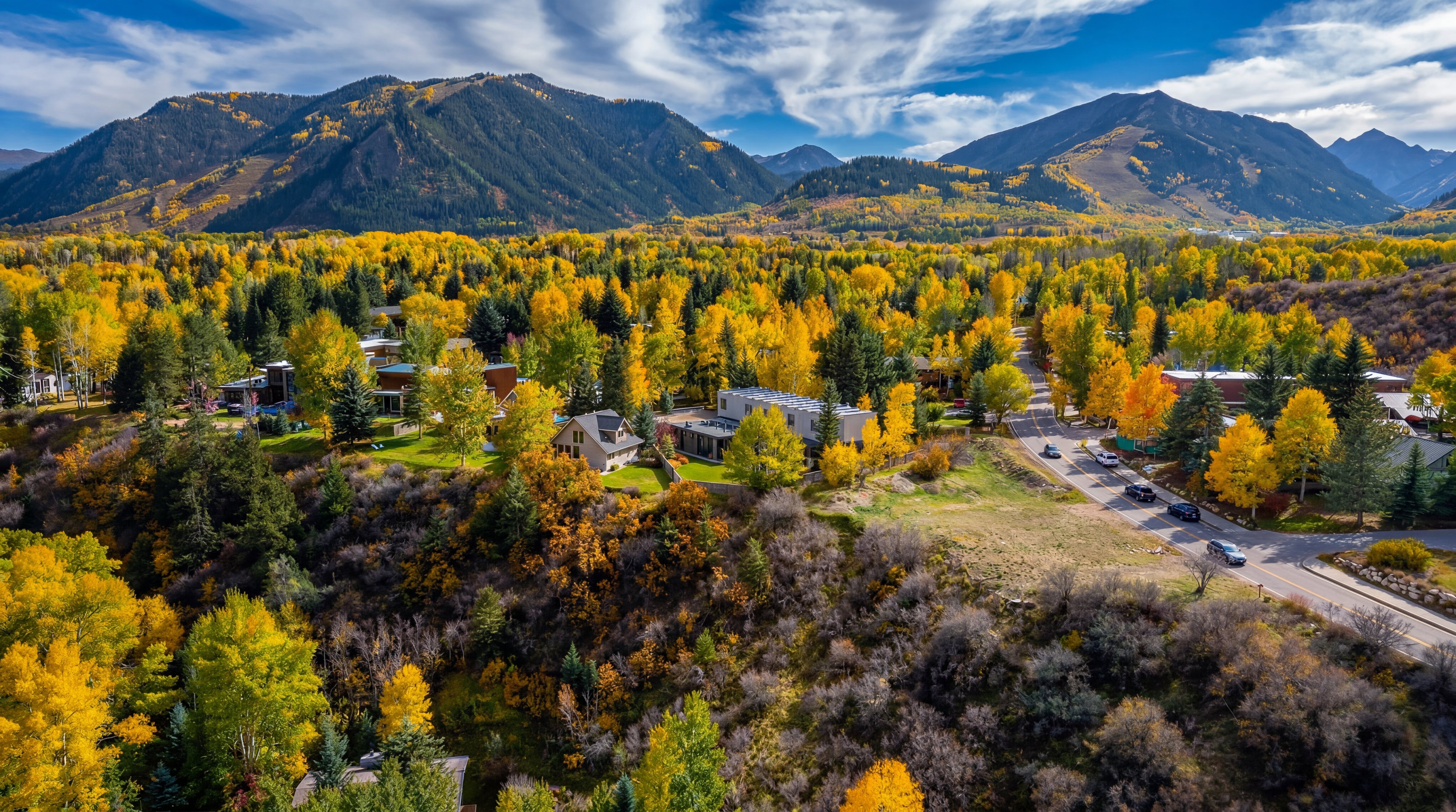 This high-angle aerial perspective captures a scenic residential neighborhood nestled within a vibrant, autumn-colored forest. The landscape is dominated by golden-yellow aspen trees and evergreen pines, set against a backdrop of majestic, rugged mountains under a bright, cloud-dappled sky. A winding road cuts through the foliage, leading toward several homes integrated into the natural, hilly terrain.