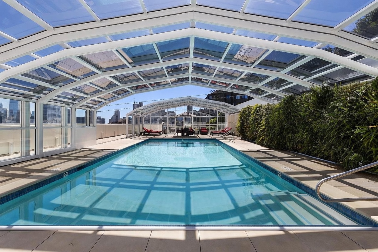 This image captures a serene indoor-outdoor swimming pool enclosed within a large, glass-paneled structure that allows for abundant natural light. The pool area features a long, rectangular basin with a tiled edge, surrounded by a paved deck and a lush vertical garden wall on one side. In the background, the structure opens up to reveal a panoramic view of a city skyline, creating a sophisticated urban oasis atmosphere.