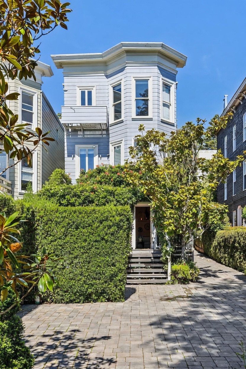 This front-facing view captures a charming, light blue multi-story home featuring a prominent bay window structure and a classic architectural style. A lush, manicured hedge provides privacy, leading to a set of stairs that ascend to the main entrance. The foreground is paved with stone pavers, complemented by a mature tree and neatly trimmed landscaping, creating a welcoming and serene curb appeal.