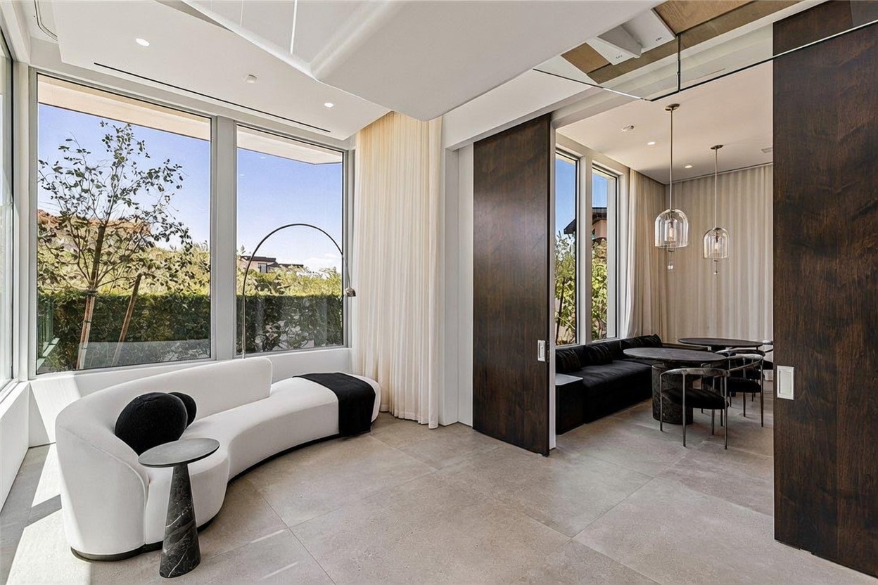 This sophisticated interior space features a curved, modern white sofa positioned near large windows that offer a view of the outdoors. To the right, dark wood sliding doors open into a dining area furnished with a round table and black seating, illuminated by elegant pendant lights. The room is finished with neutral-toned large-format floor tiles, creating a seamless and contemporary aesthetic.