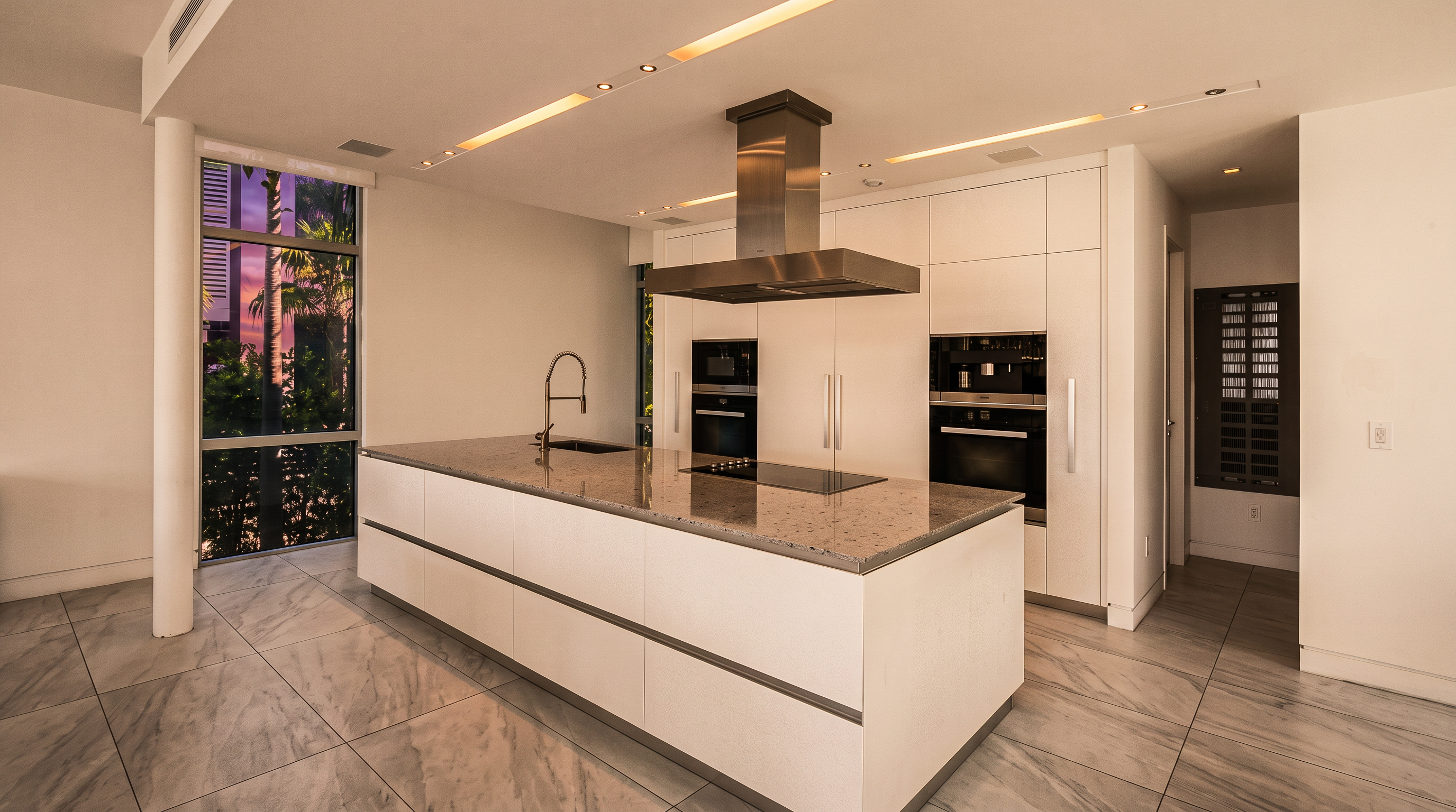 This modern, minimalist kitchen features a large central island with a stone countertop and an integrated cooktop beneath a stainless steel range hood. The space is defined by sleek, white cabinetry that extends to the ceiling, complemented by light-colored tile flooring and large windows that provide ample natural light. The clean lines and neutral color palette create a bright, airy, and contemporary atmosphere.