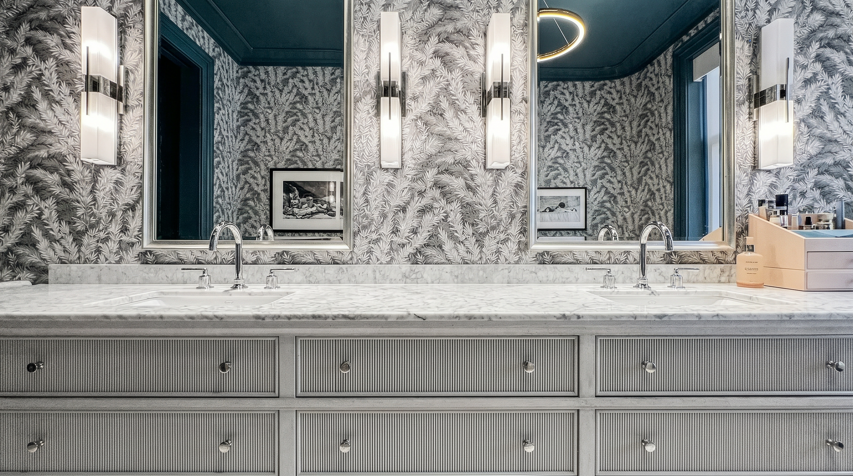 This elegant primary bathroom features a long, custom-built vanity with textured drawer fronts and a light-toned stone countertop. The space is defined by bold, monochromatic botanical wallpaper and a dark-painted ceiling, complemented by sleek wall-mounted light fixtures and framed mirrors. The perspective is a wide-angle shot capturing the full length of the vanity, emphasizing the sophisticated and high-end design aesthetic.