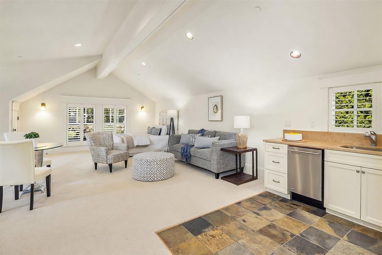 This spacious, open-concept studio suite features a vaulted ceiling and a seamless blend of living, sleeping, and kitchenette areas. The room is anchored by a comfortable gray sofa, a patterned ottoman, and a cozy bed, all set against a neutral color palette with soft carpeting. A small dining area and a functional kitchenette with slate-tiled flooring complete this versatile and inviting living space.