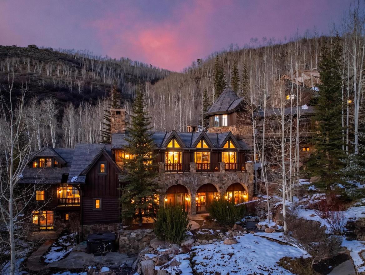 This stunning mountain estate is captured at twilight, showcasing a multi-level architectural design with a blend of dark wood siding and natural stone masonry. The home features a series of illuminated windows, a covered stone-arched patio on the lower level, and a spacious balcony above, all set against a backdrop of dense aspen trees and a snow-dusted landscape. The warm glow from the interior lights contrasts beautifully with the soft purple and pink hues of the sunset sky, creating an inviting and luxurious atmosphere.