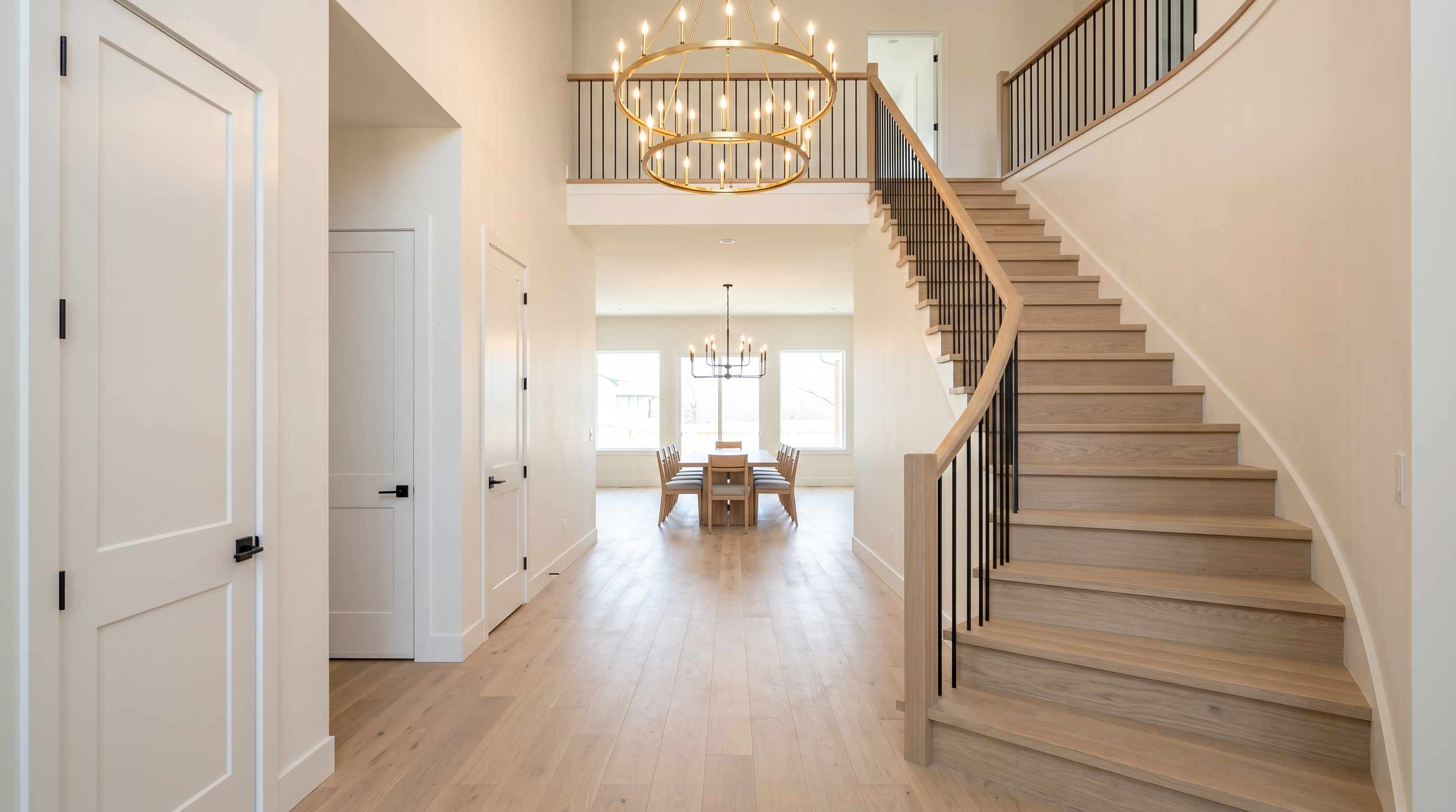 This grand entryway features a sweeping wooden staircase with modern black railings and light-toned hardwood flooring that carries into a formal dining area. The space is illuminated by a striking tiered brass chandelier, creating a bright, open, and high-end aesthetic. The neutral color palette and clean lines establish an elegant, modern architectural character throughout the home.