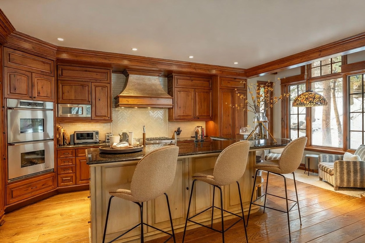 This warm and inviting kitchen features rich, dark wood cabinetry paired with a large central island topped with dark stone countertops. The space is accented by a custom metal range hood, a herringbone-patterned backsplash, and three upholstered bar stools. Large windows to the right allow for ample natural light, creating a bright and comfortable atmosphere perfect for cooking and entertaining.