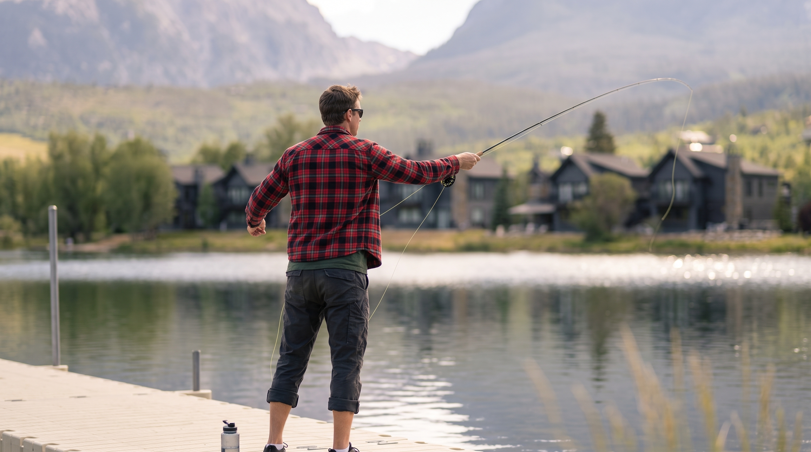 This image captures a serene lakeside scene, highlighting a man fly-fishing from a floating dock in a tranquil, resort-like community setting. The background features upscale, modern mountain-style homes nestled near the water, framed by majestic, hazy mountain peaks. This picturesque environment suggests high-end recreational amenities and an active, nature-oriented lifestyle typical of premium residential developments.