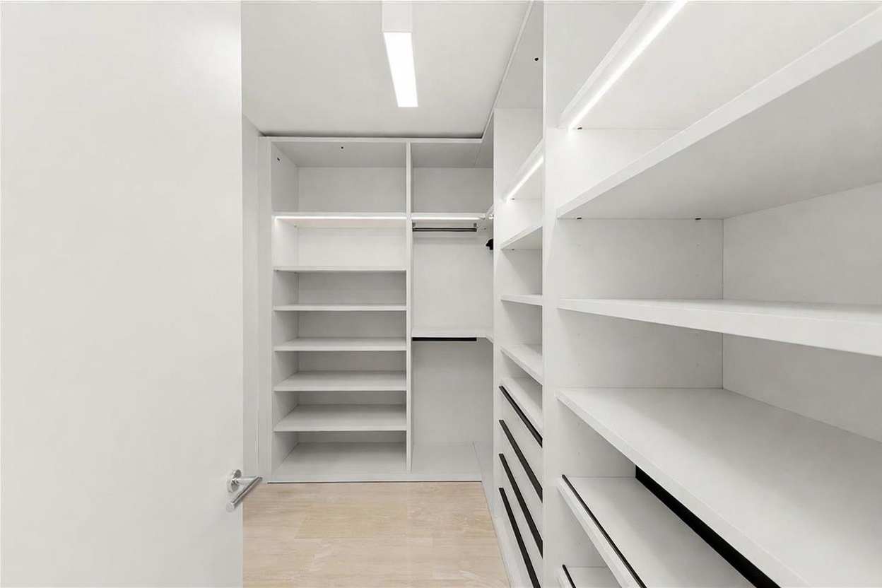 This modern walk-in closet features crisp white custom cabinetry with integrated LED lighting along the shelves. The space is organized with a mix of open shelving, hanging rods, and a series of drawers, all set against a clean, minimalist aesthetic. The light-toned flooring complements the bright, airy atmosphere of the room.