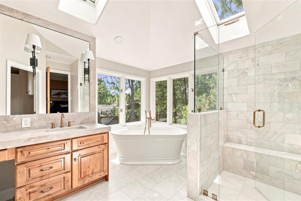 This bright primary bathroom features a freestanding soaking tub positioned beneath large windows that offer a view of the surrounding trees. To the left, a wooden vanity with a stone countertop and dual wall-mounted light fixtures provides a warm contrast to the light-toned stone flooring and shower tile. A glass-enclosed walk-in shower with a built-in bench sits to the right, while a skylight above adds natural illumination to the airy, high-ceilinged space.
