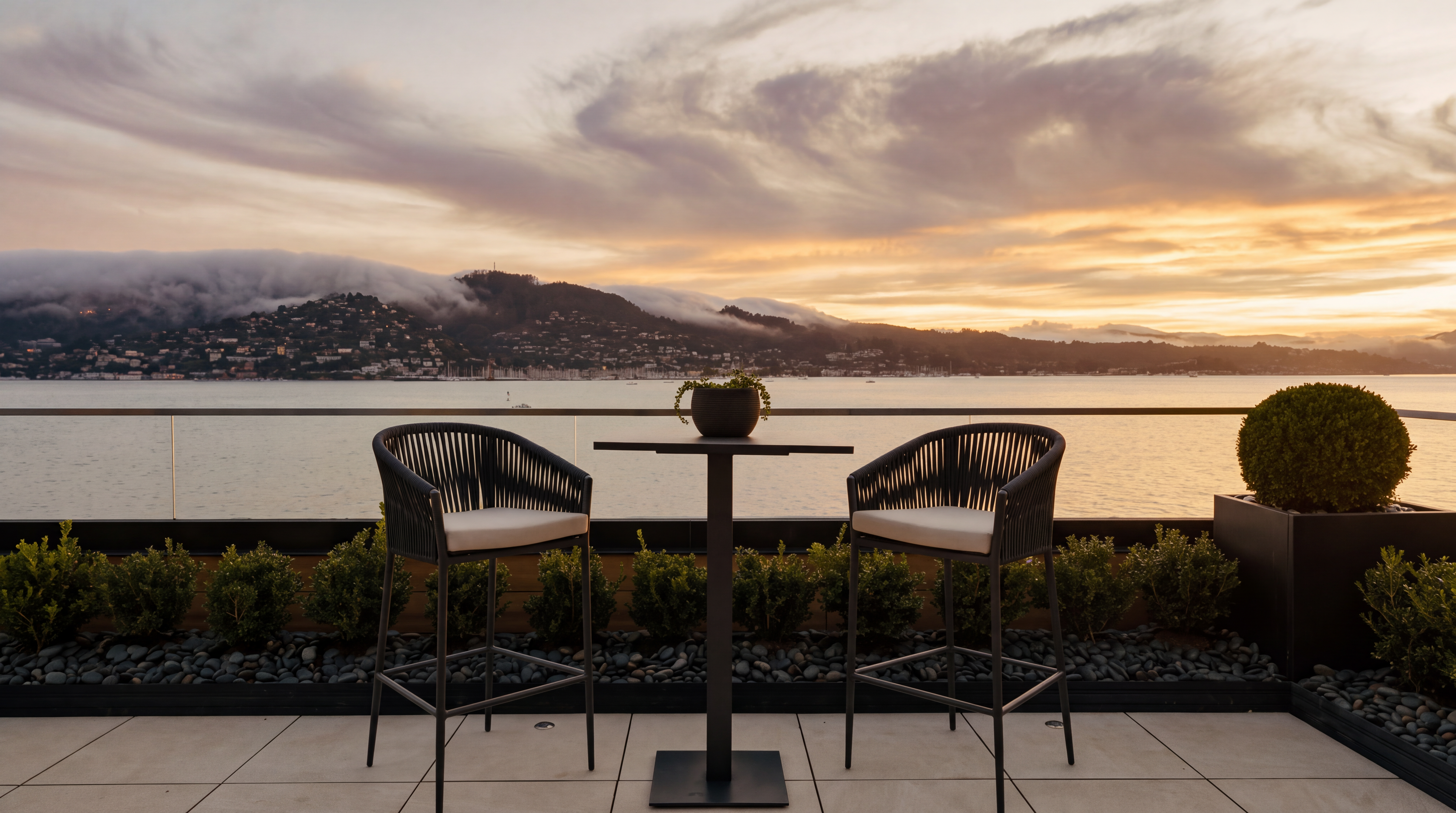 This elevated balcony offers a stunning, unobstructed view of a bay at sunset, with dramatic clouds hovering over the distant hills. The space is furnished with two modern, dark-toned woven chairs and a matching bistro table, creating an intimate setting for enjoying the scenery. A sleek glass railing ensures the view remains the focal point, while a neat row of small shrubs and a potted plant add a touch of greenery to the tiled terrace.