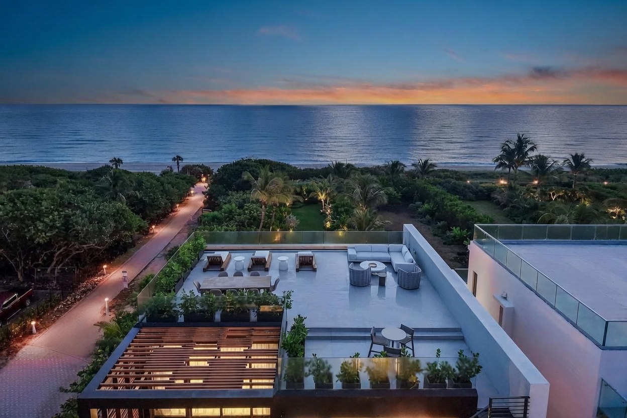 This high-angle aerial view captures a luxurious rooftop terrace overlooking a serene ocean horizon at sunset. The deck is furnished with a modern sectional, lounge chairs, and a dining set, all arranged to maximize the panoramic coastal views. Below the terrace, lush tropical landscaping transitions into a private path leading toward the sandy beach, creating an atmosphere of exclusive seaside living.