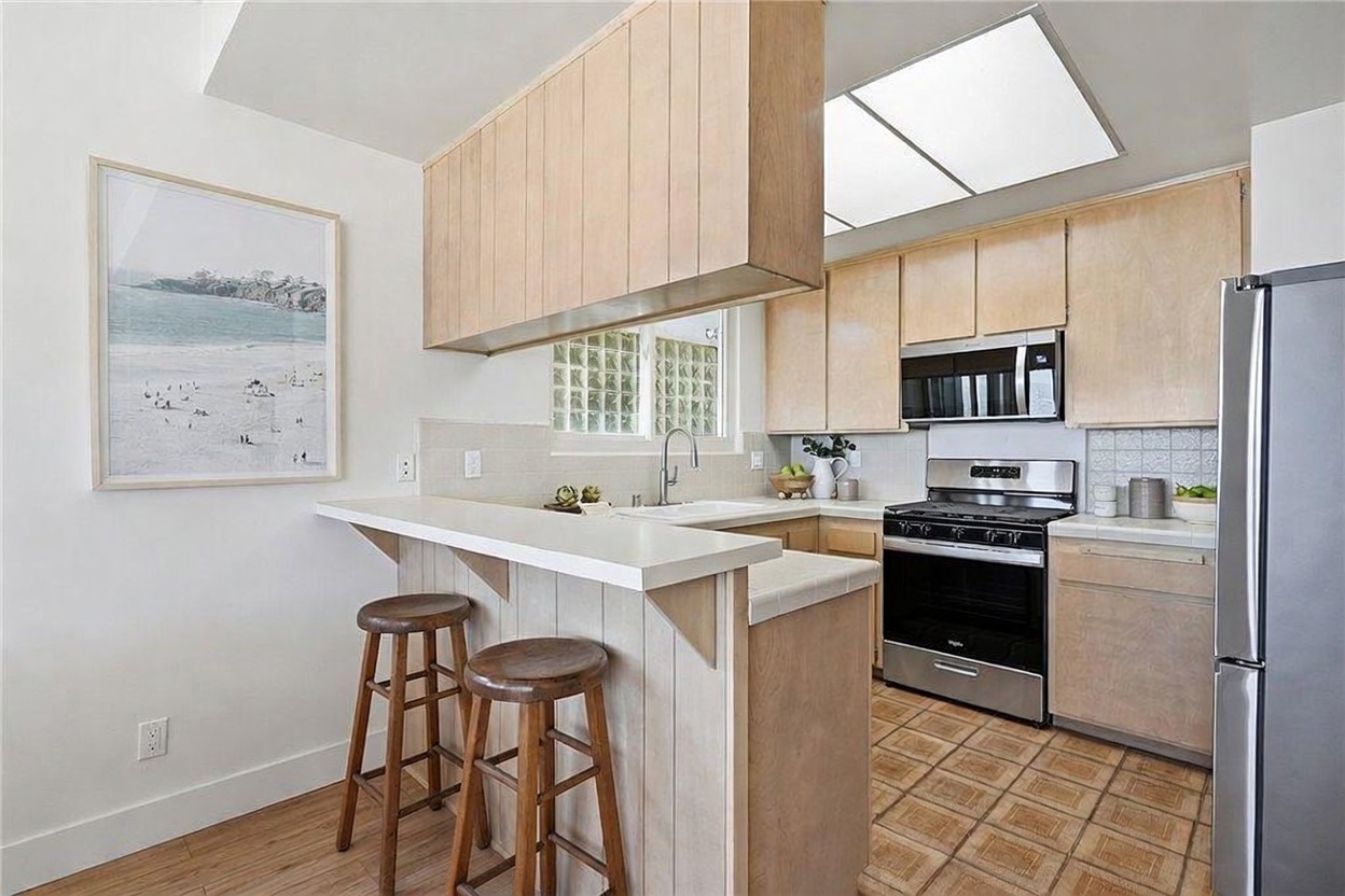 This kitchen features light wood cabinetry, a breakfast bar with two wooden stools, and a stainless steel stove and microwave. The space includes a tiled floor, a sink positioned under a window with glass block accents, and a recessed ceiling light. The overall aesthetic is functional and bright, offering a clear view of the cooking and dining areas from a slightly elevated perspective.