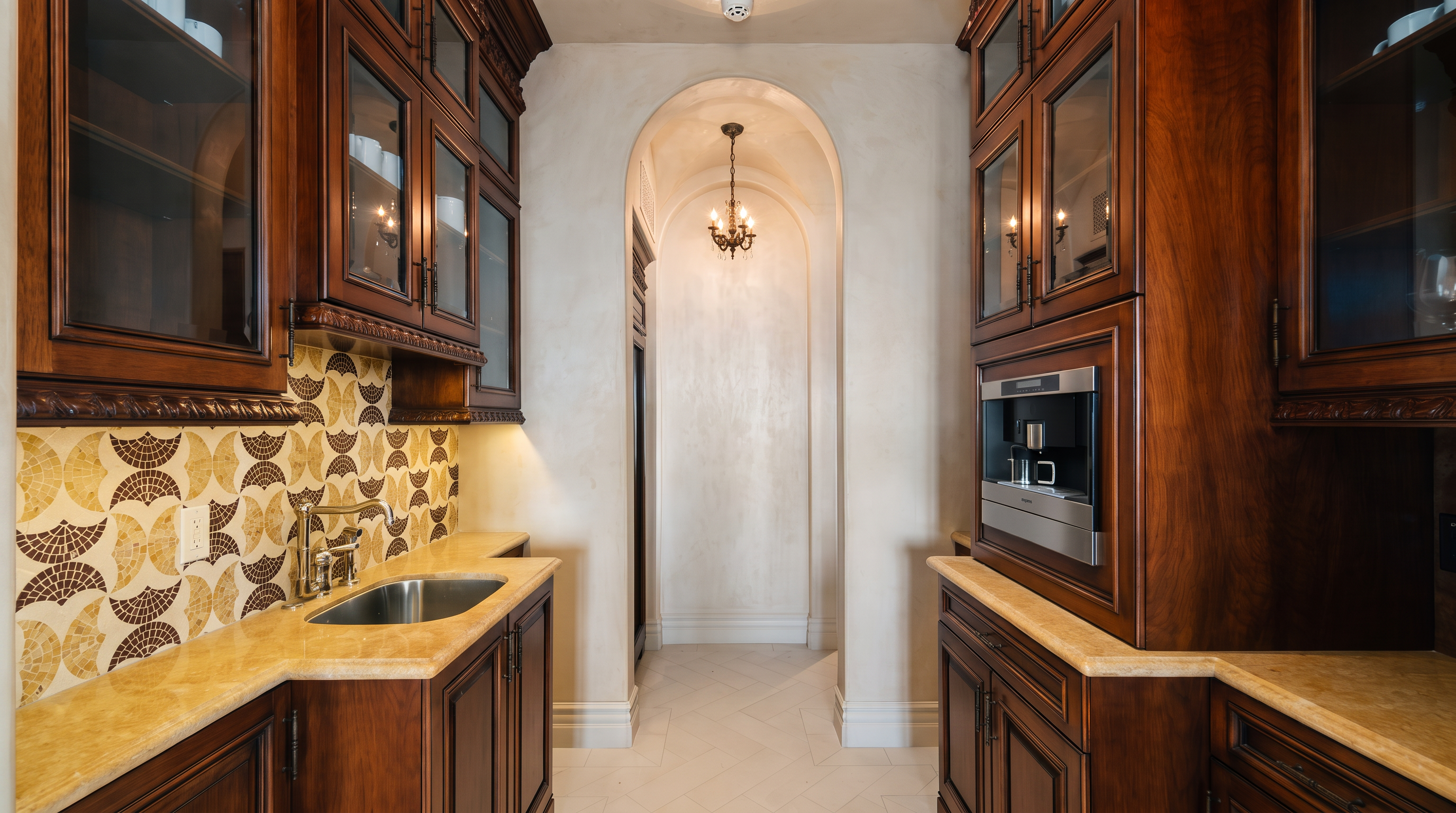 This elegant butler's pantry features rich, dark wood cabinetry with glass-front upper doors and a built-in coffee machine. A light-colored stone countertop complements the intricate, patterned tile backsplash and the undermount sink, while an arched walkway leads to a secondary space illuminated by a small chandelier. The perspective is centered, highlighting the symmetrical layout and the sophisticated, traditional design of the transition area.