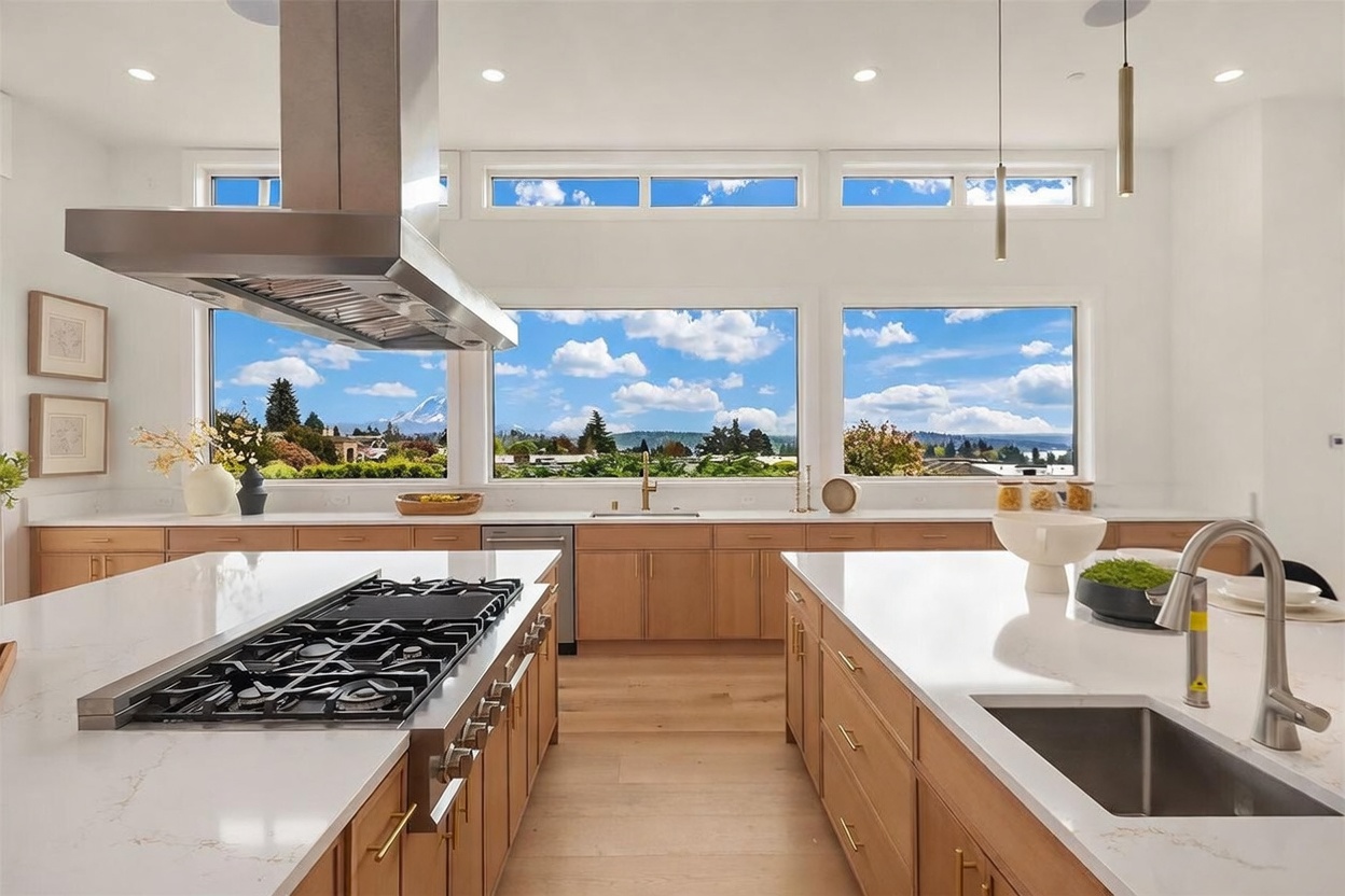 This bright, modern kitchen features light wood cabinetry paired with white countertops and a large central island equipped with a gas cooktop. A stainless steel range hood hangs above the island, while a double-window setup provides an expansive view of the outdoors. The space is illuminated by recessed lighting and sleek pendant lights, creating a clean and airy atmosphere.