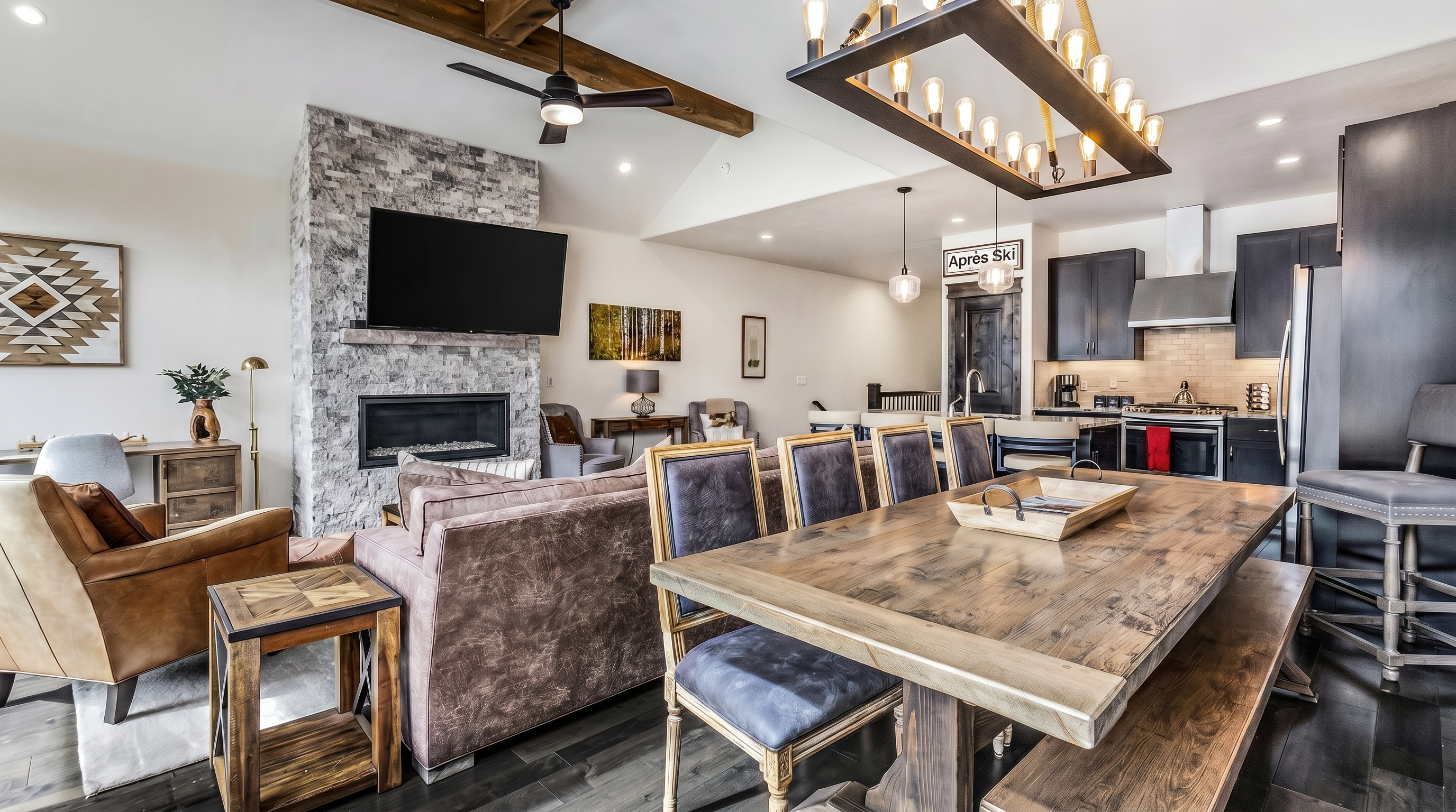 This open-concept living and dining area showcases a rustic-modern aesthetic with exposed wooden ceiling beams, a stone-clad fireplace, and dark hardwood flooring. The space features a large wooden dining table, a cozy leather armchair, and a comfortable sofa, all illuminated by a striking industrial-style chandelier. The perspective captures the seamless flow between the living room and the kitchen, highlighting the home's inviting and spacious atmosphere.