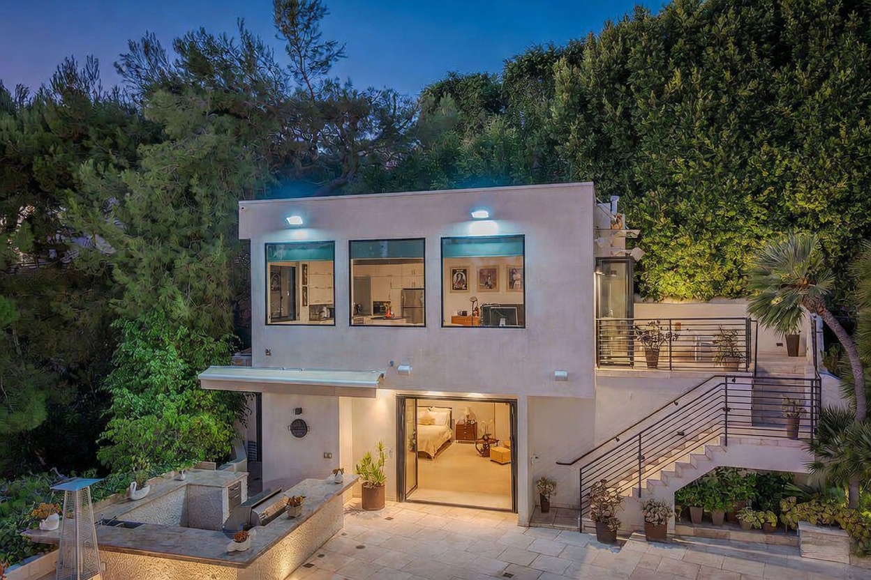 This contemporary multi-level home features a clean, light-colored stucco facade with large windows that provide a view into the upper-level living spaces. The lower level opens directly onto a stone-paved patio equipped with an outdoor kitchen and bar area, while an external staircase leads up to a private balcony. The property is nestled against a lush, dense hillside, creating a secluded and tranquil atmosphere at dusk.