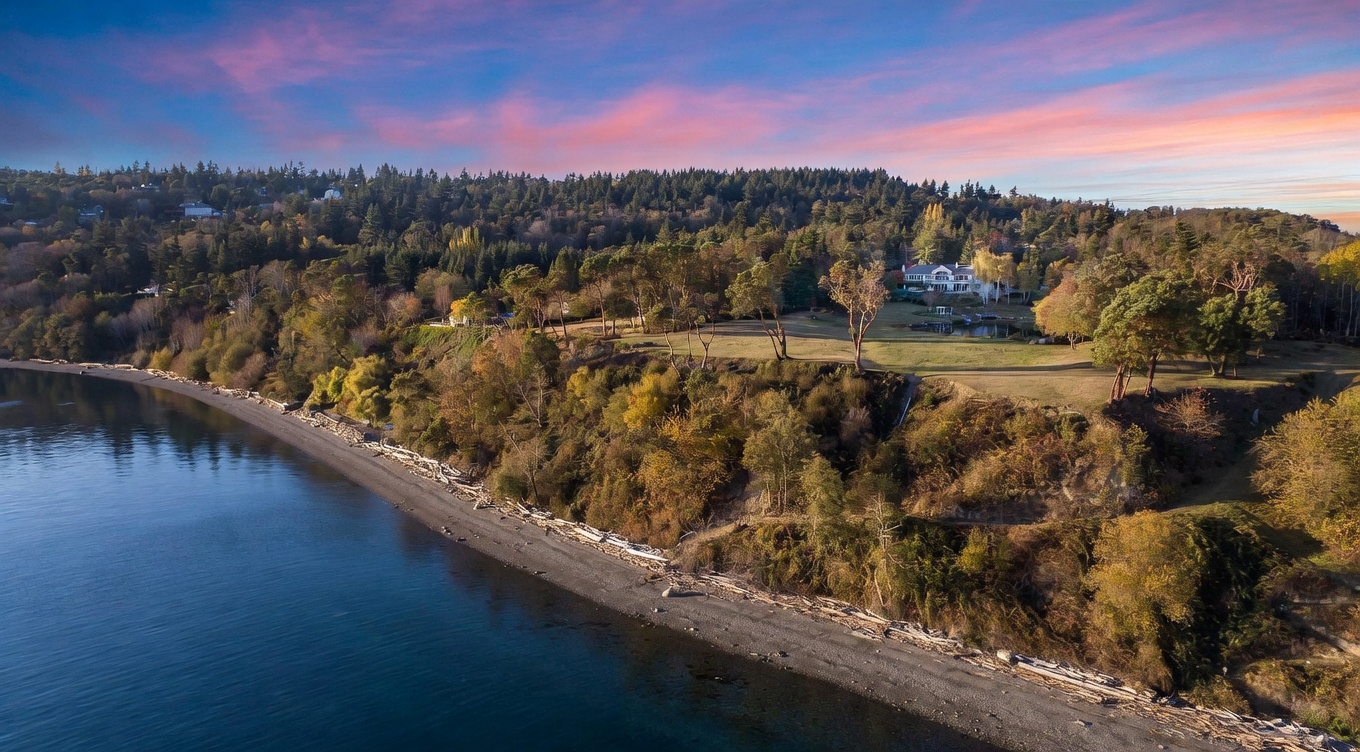 This wide-angle aerial perspective captures a sprawling coastal estate perched atop a lush, forested bluff overlooking a serene beach. The property features a large residence nestled among mature trees, expansive green lawns, and a small pond, all set against a dramatic sunset sky. The composition emphasizes the property's secluded location, natural topography, and direct access to the shoreline.