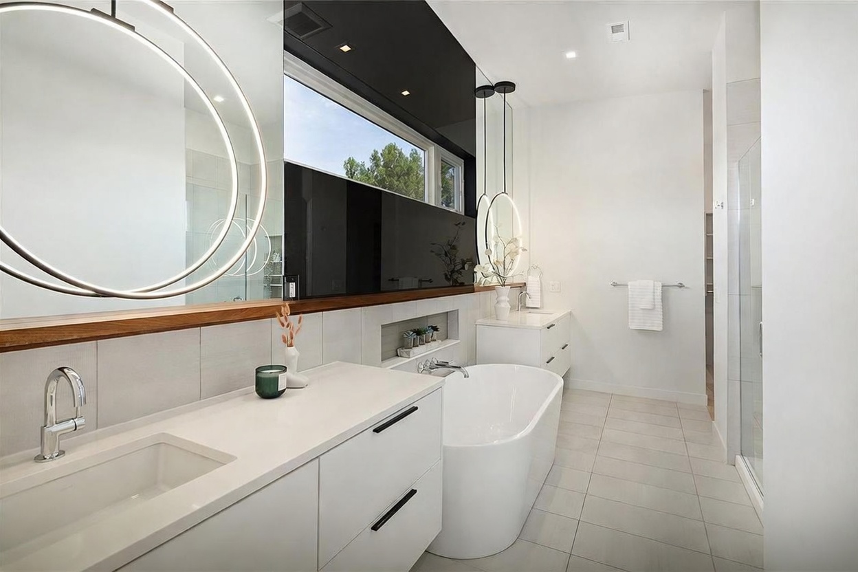 This modern primary bathroom features a sleek, minimalist aesthetic with a white double vanity, a freestanding soaking tub, and a large, circular backlit mirror. A horizontal window above the tub provides natural light, while a recessed niche offers convenient storage. The space is finished with light-toned tile flooring and a glass-enclosed shower, creating a bright and airy atmosphere.