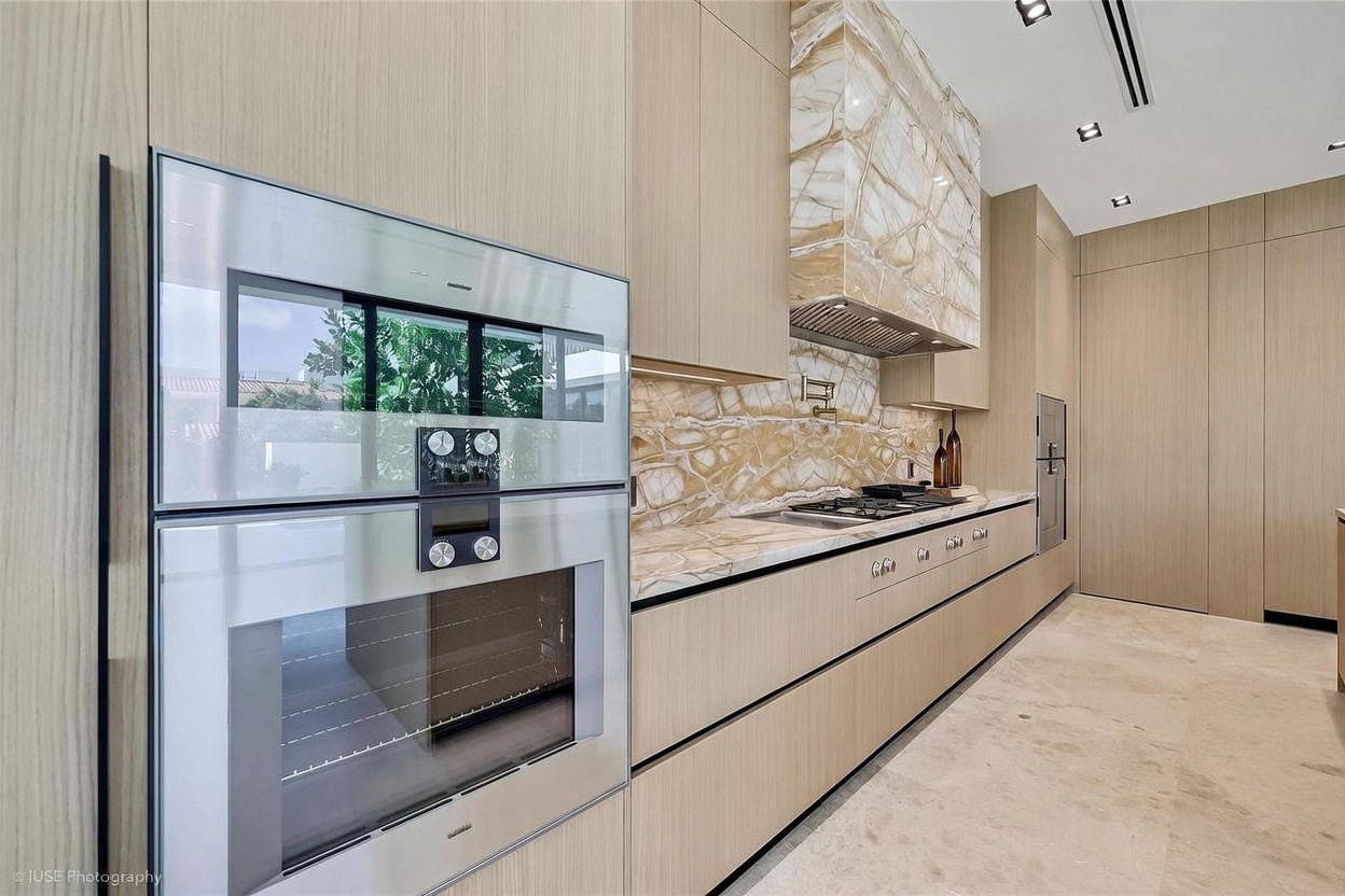 This sleek, modern kitchen features light wood cabinetry and a striking stone backsplash that extends up to a matching stone-clad range hood. The space is equipped with high-end stainless steel appliances, including a built-in oven and a gas cooktop set into a stone countertop. The perspective captures the linear flow of the cabinetry and the polished stone flooring, creating a sophisticated and minimalist aesthetic.